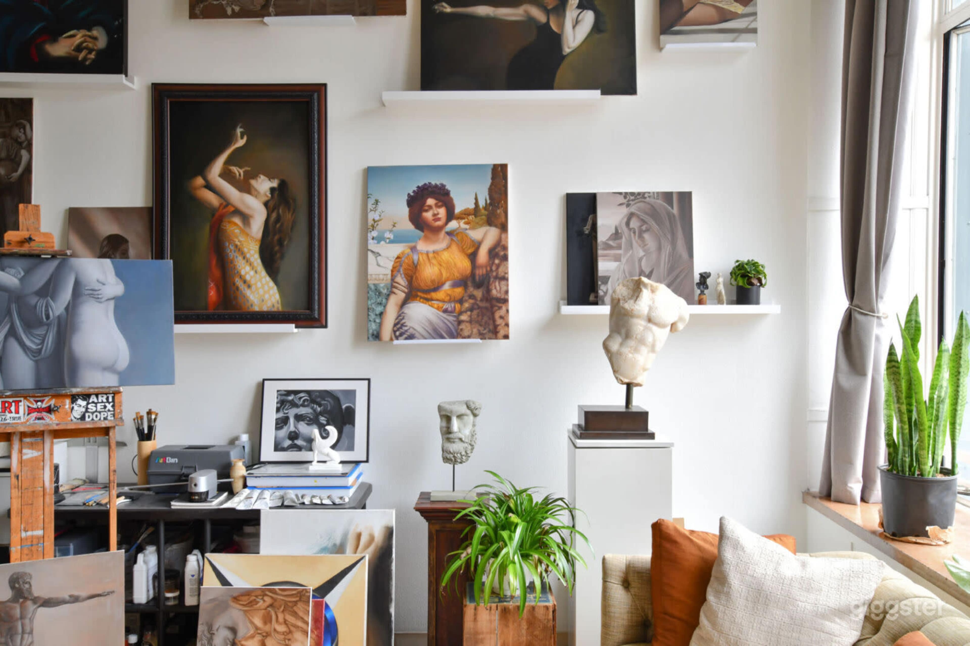 The image depicts an artist's studio featuring various paintings on the walls, a sculpture on a pedestal, and art supplies on a table.