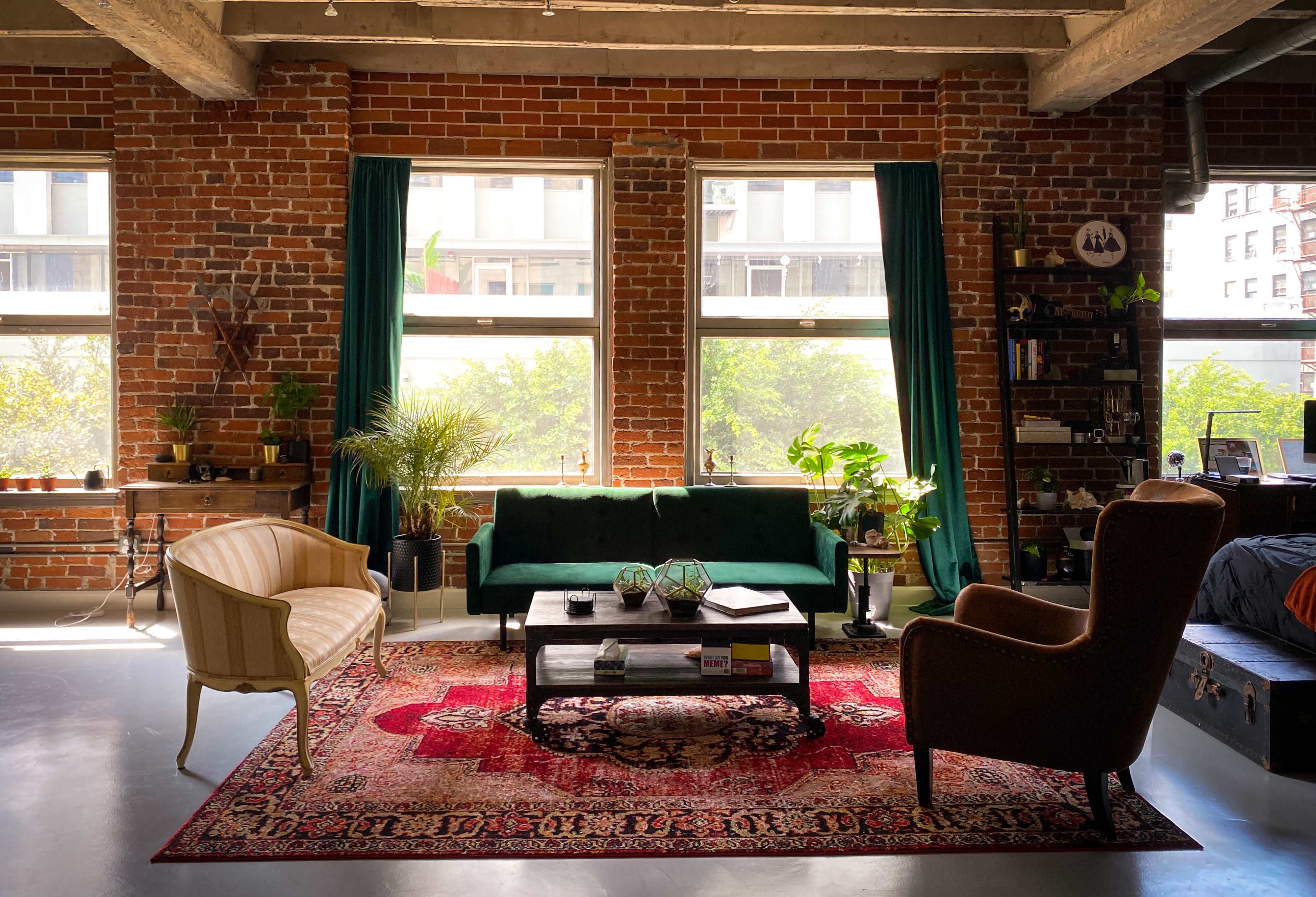 The image shows a cozy living room with a brick wall, large windows, a green sofa, an armchair, a coffee table, a patterned rug, and various indoor plants.