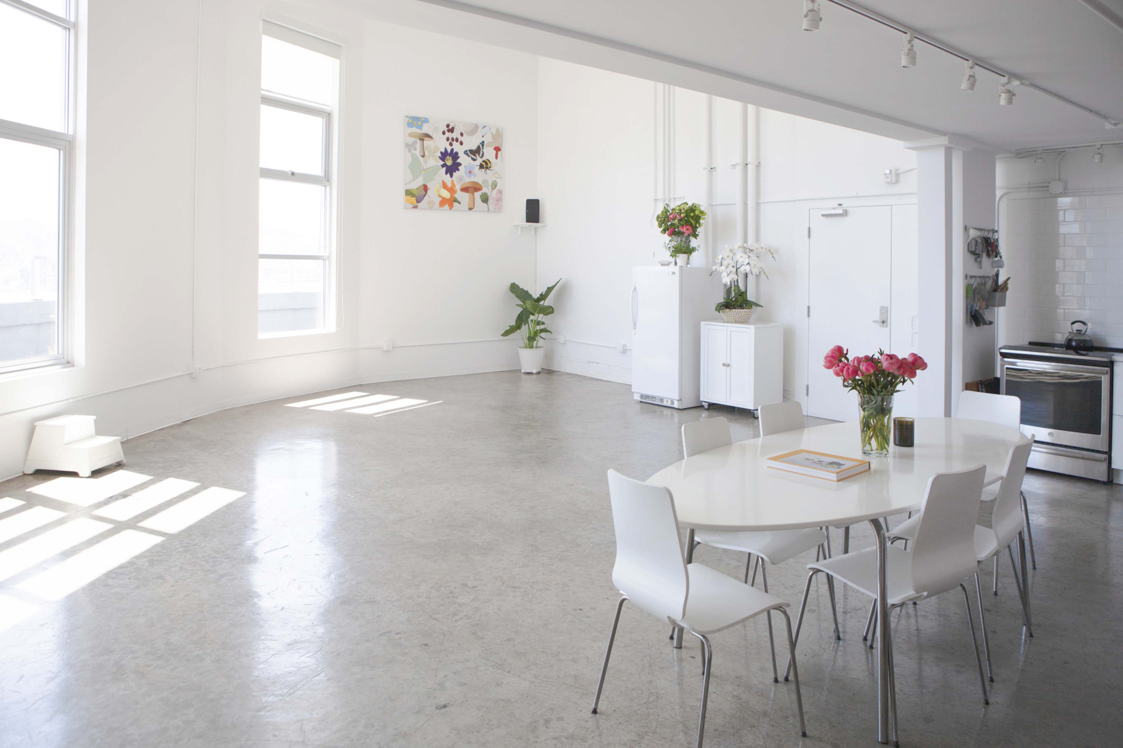 The image shows a bright, spacious interior featuring a circular dining table with white chairs, a kitchen area, and large windows allowing natural light to fill the room.