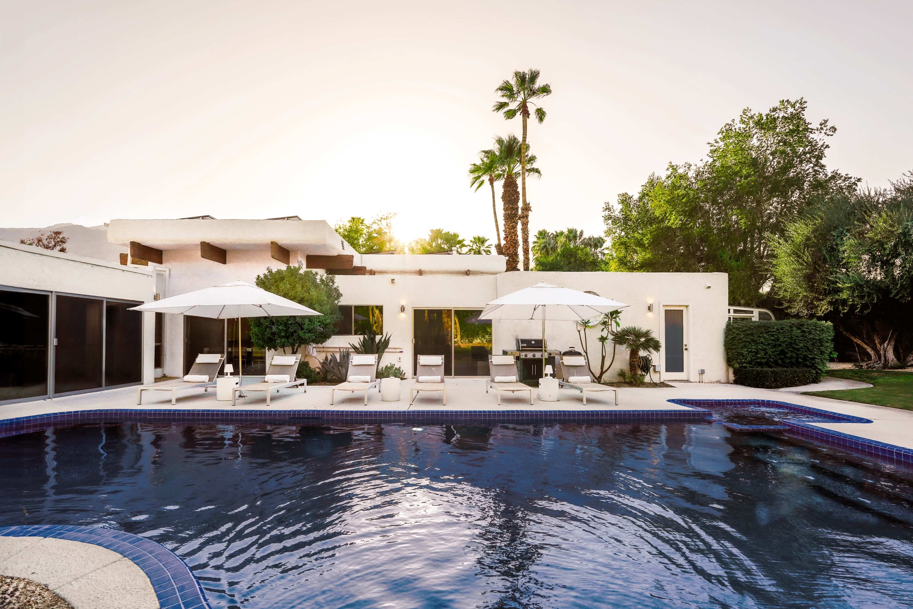 A modern house with a pool is surrounded by palm trees and lounge chairs, with large umbrellas providing shade.