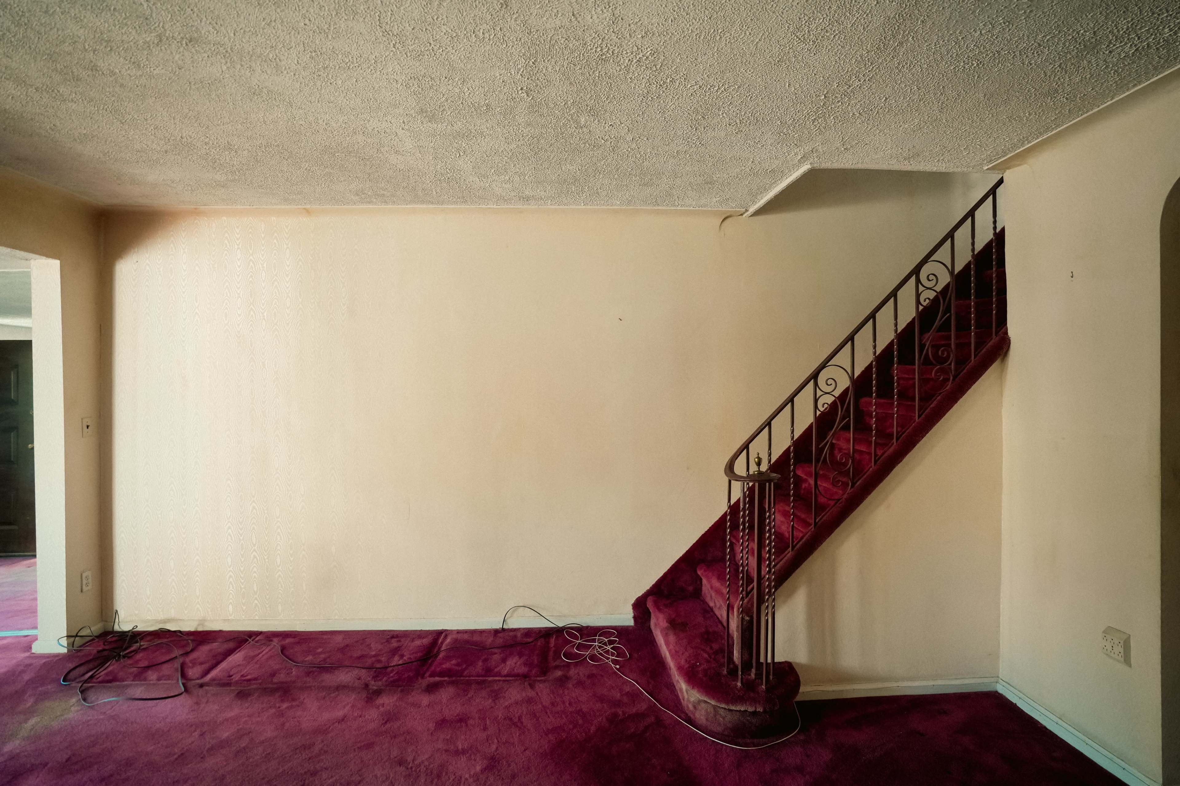 The image features an empty room with a maroon carpet and a staircase against a light-colored wall.