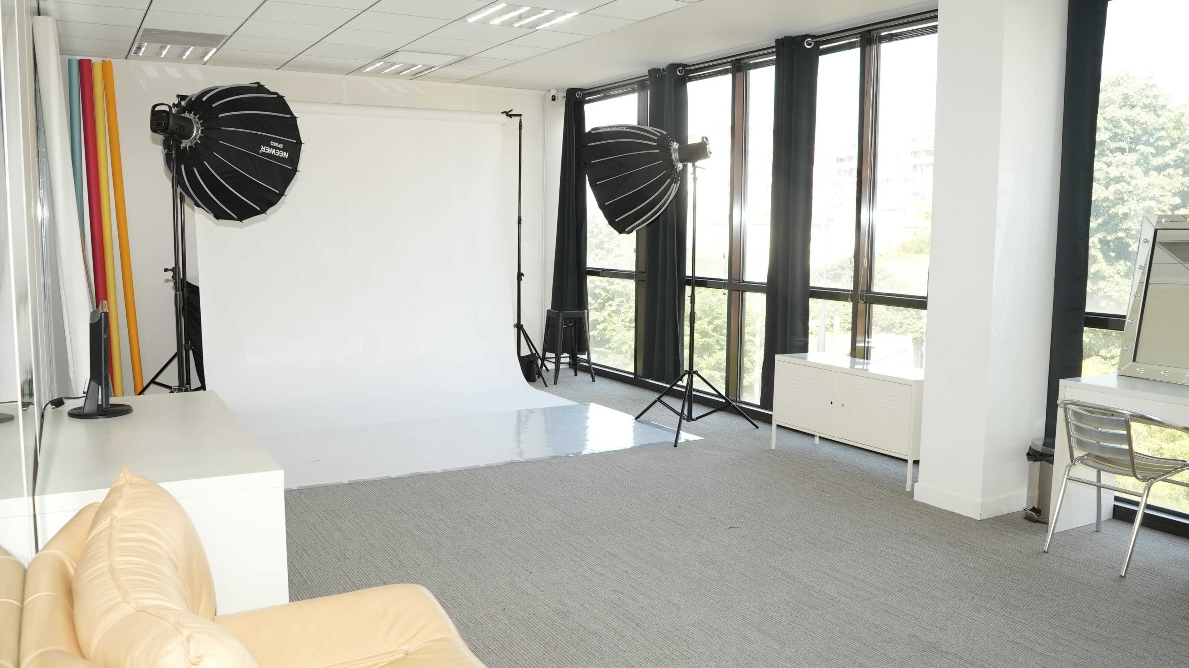 The image shows a well-lit photography studio featuring a white backdrop, two large softbox lights, and a couch in a modern space with large windows.
