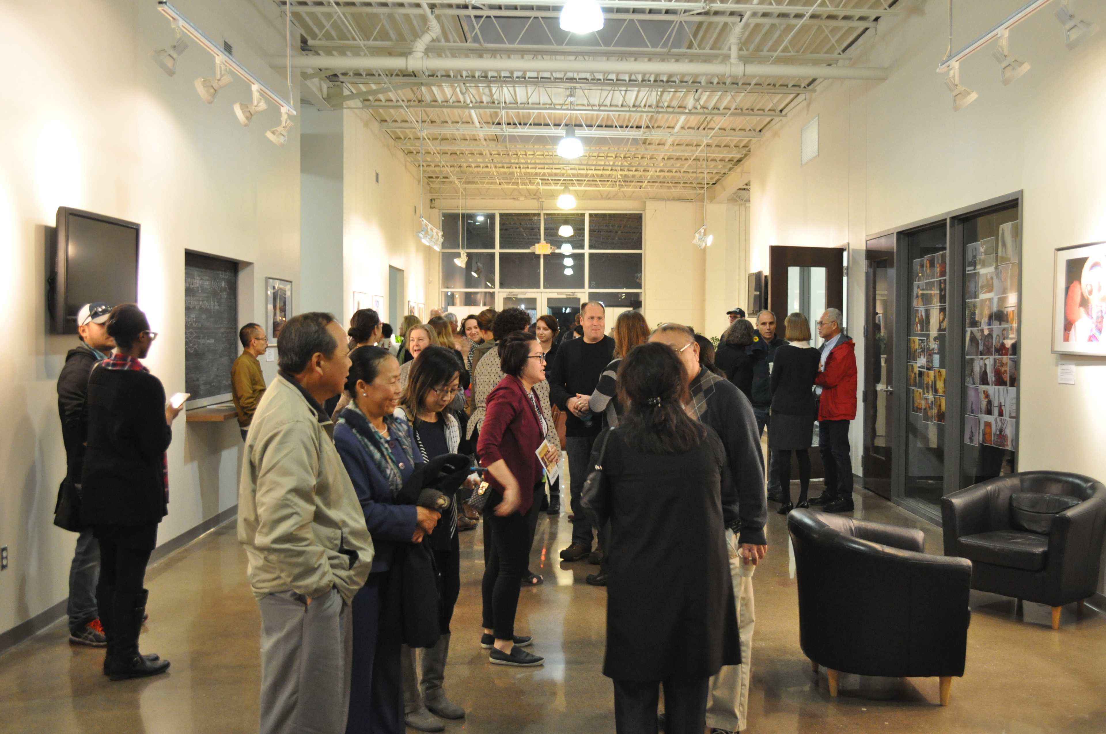 A diverse group of people is gathered in an art gallery, engaging in conversation and viewing photographs displayed on the walls.