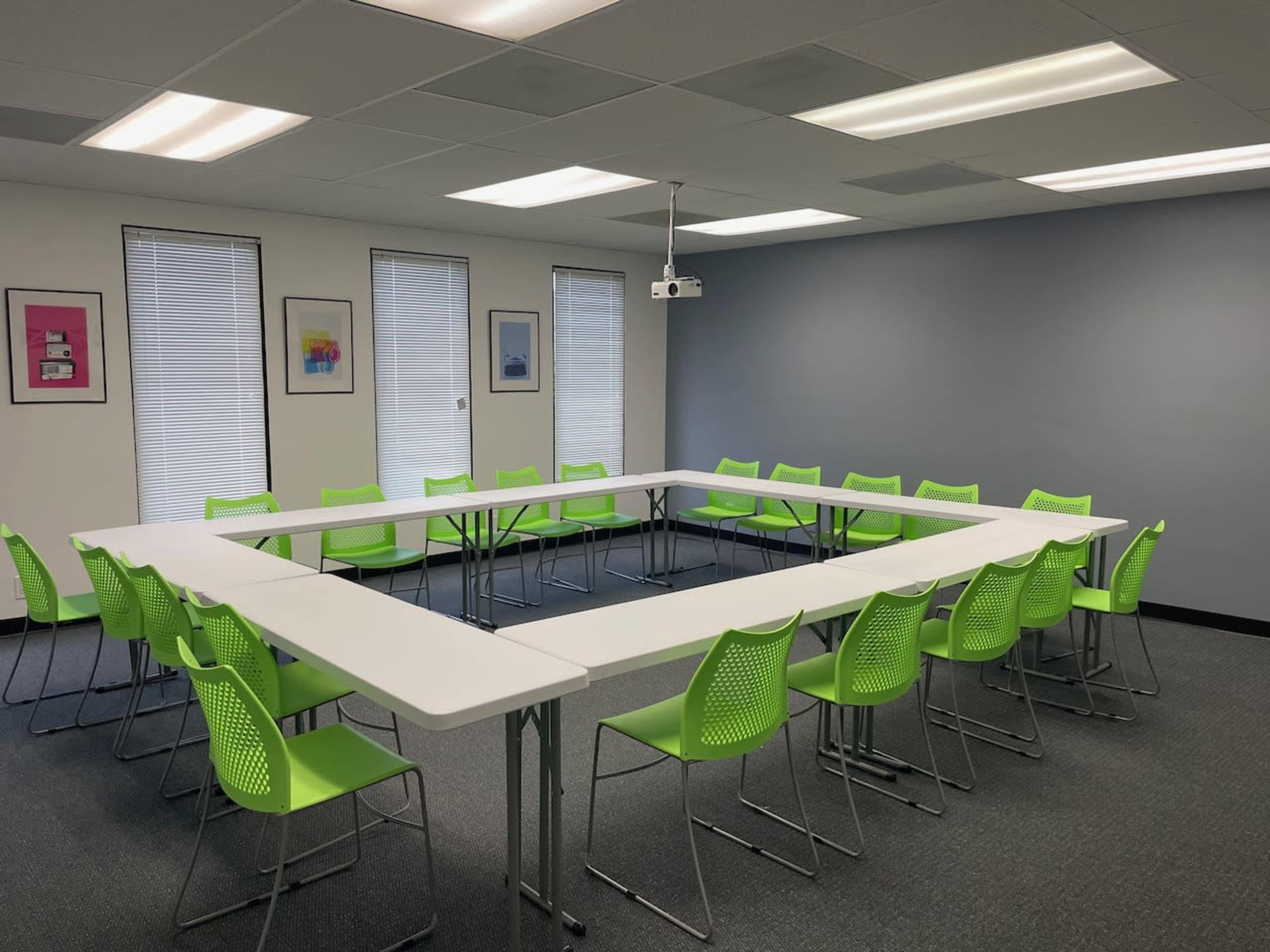 A conference room features a triangular arrangement of white tables surrounded by green chairs, with windows and artwork on the walls.