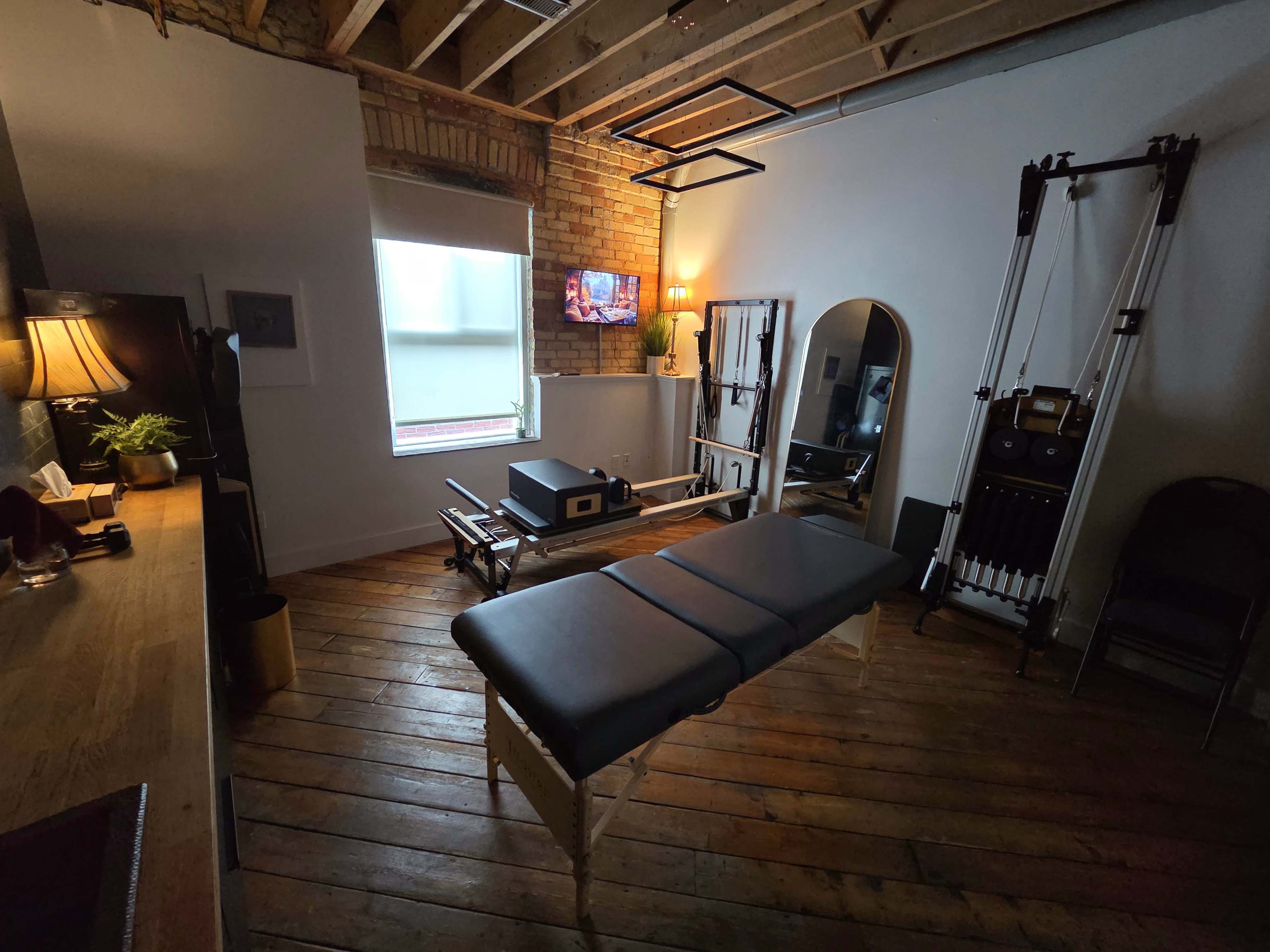 The image shows a small therapy or treatment room featuring a massage table, fitness equipment, a mirror, and a TV mounted on the wall.