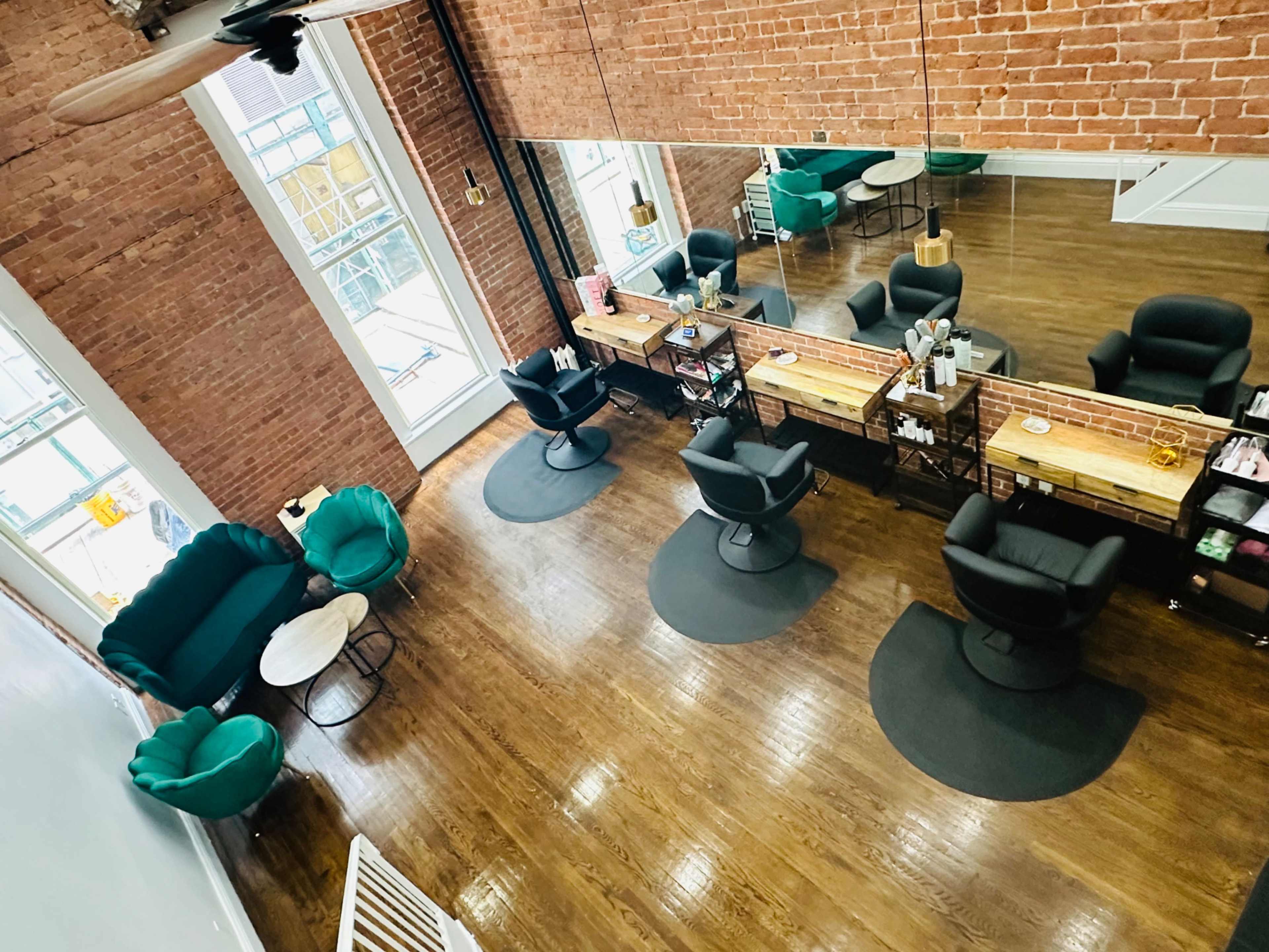 The image shows a bright salon interior with brick walls, mirrored mirrors, a wooden floor, and two types of seating arrangements.