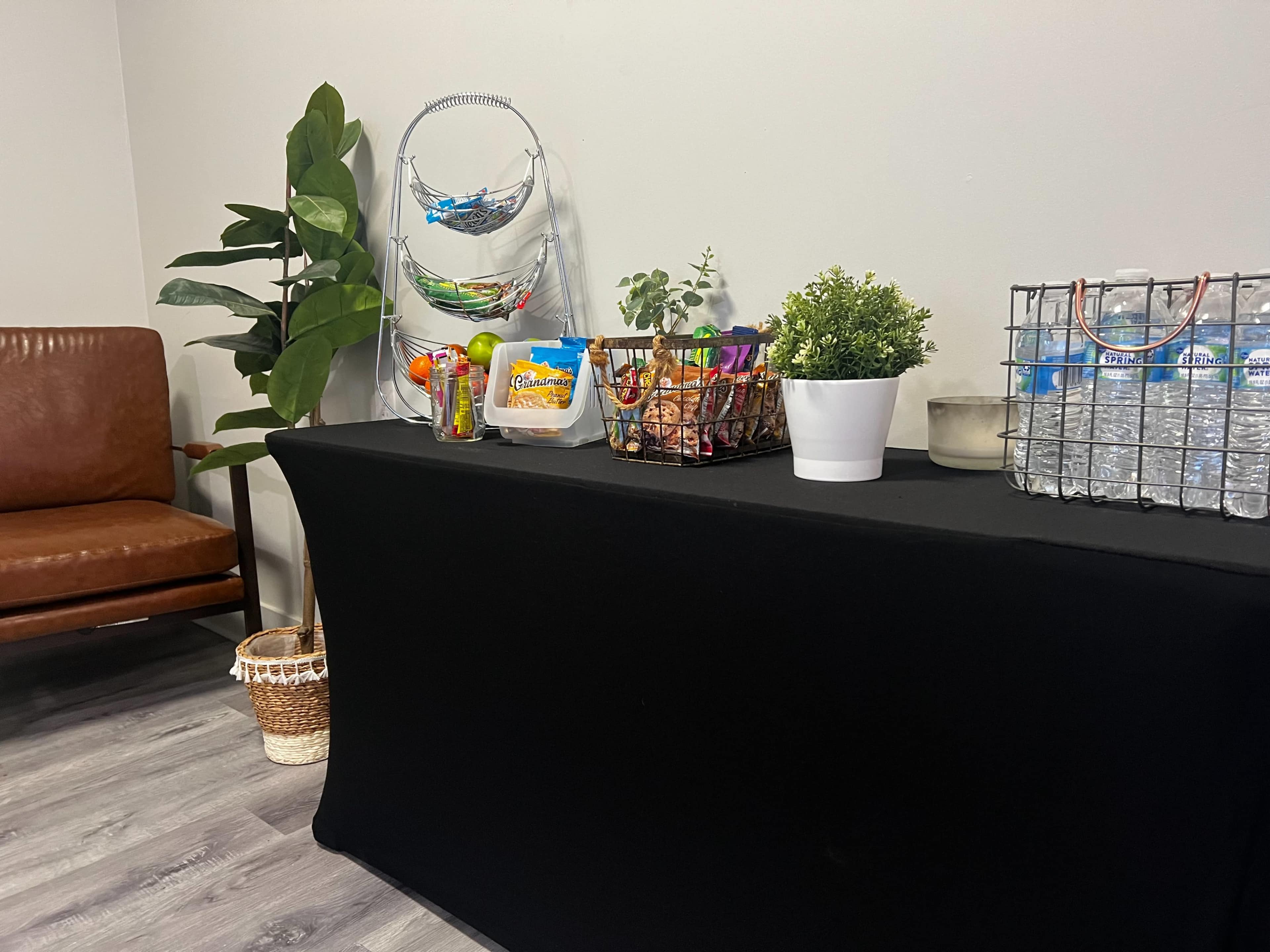 A black table displays a variety of snacks and drinks, accompanied by a potted plant and a chair nearby.