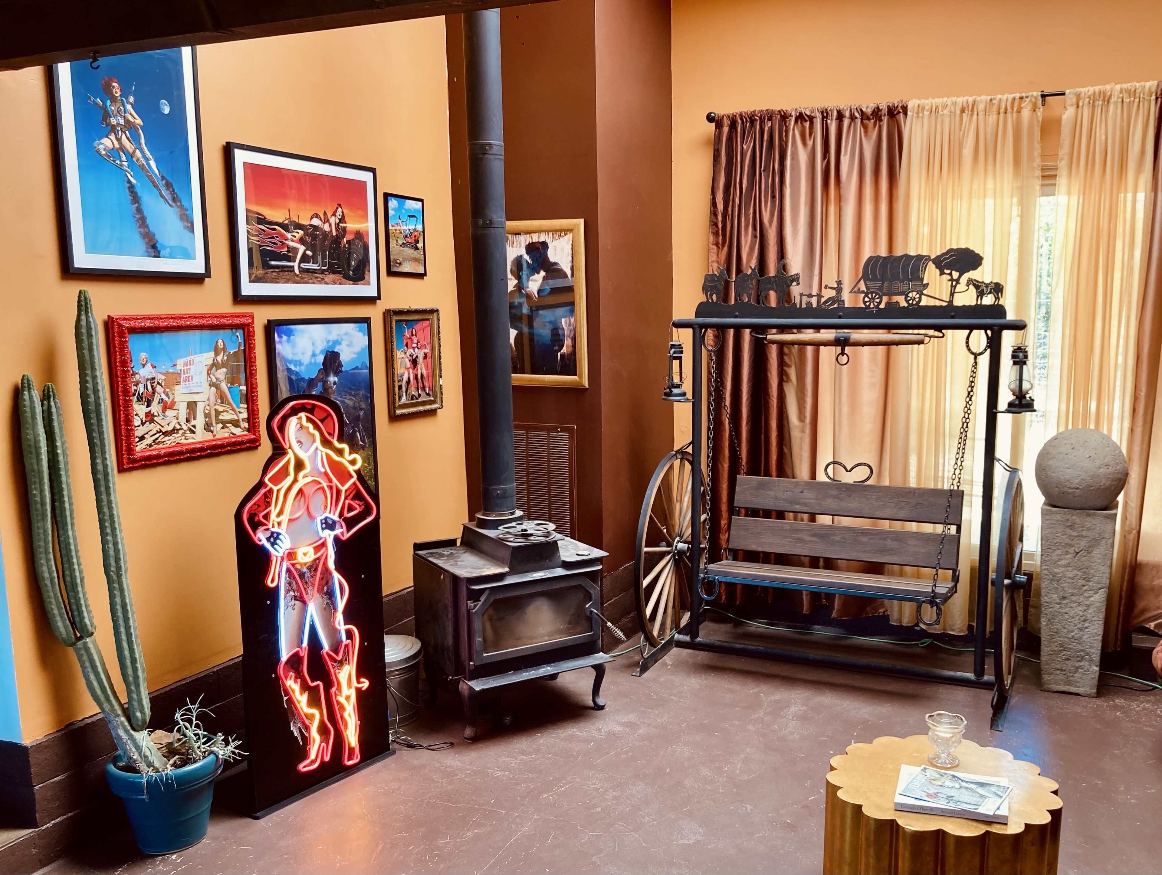 The scene features a corner of a room with a neon figure of a woman, a wooden swing, a wood stove, framed photographs on the walls, a large round stone, and a cactus.