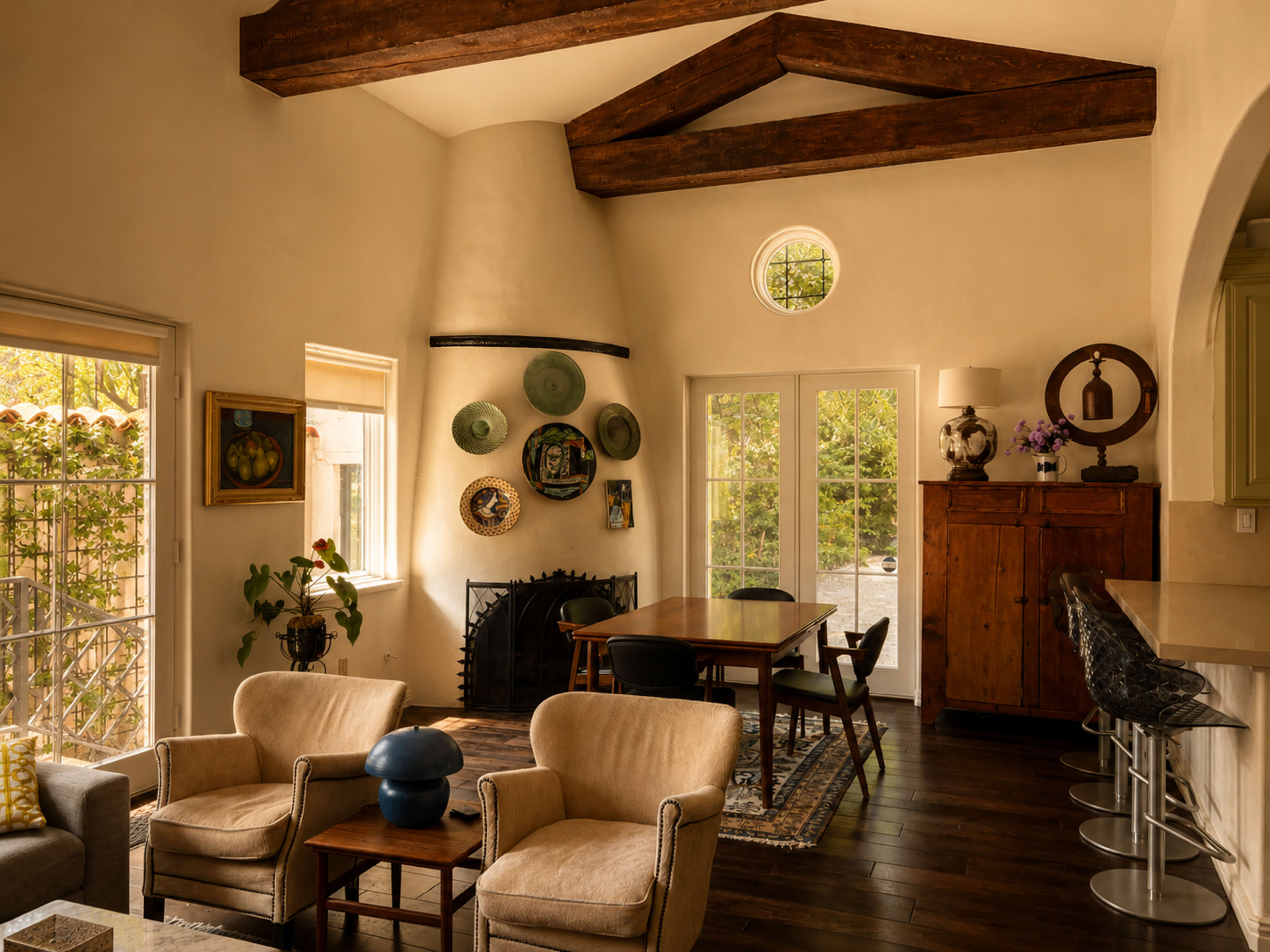 The image shows a cozy living space with a fireplace, wooden beams, and a dining area featuring a table and chairs, surrounded by decorative items on the walls.