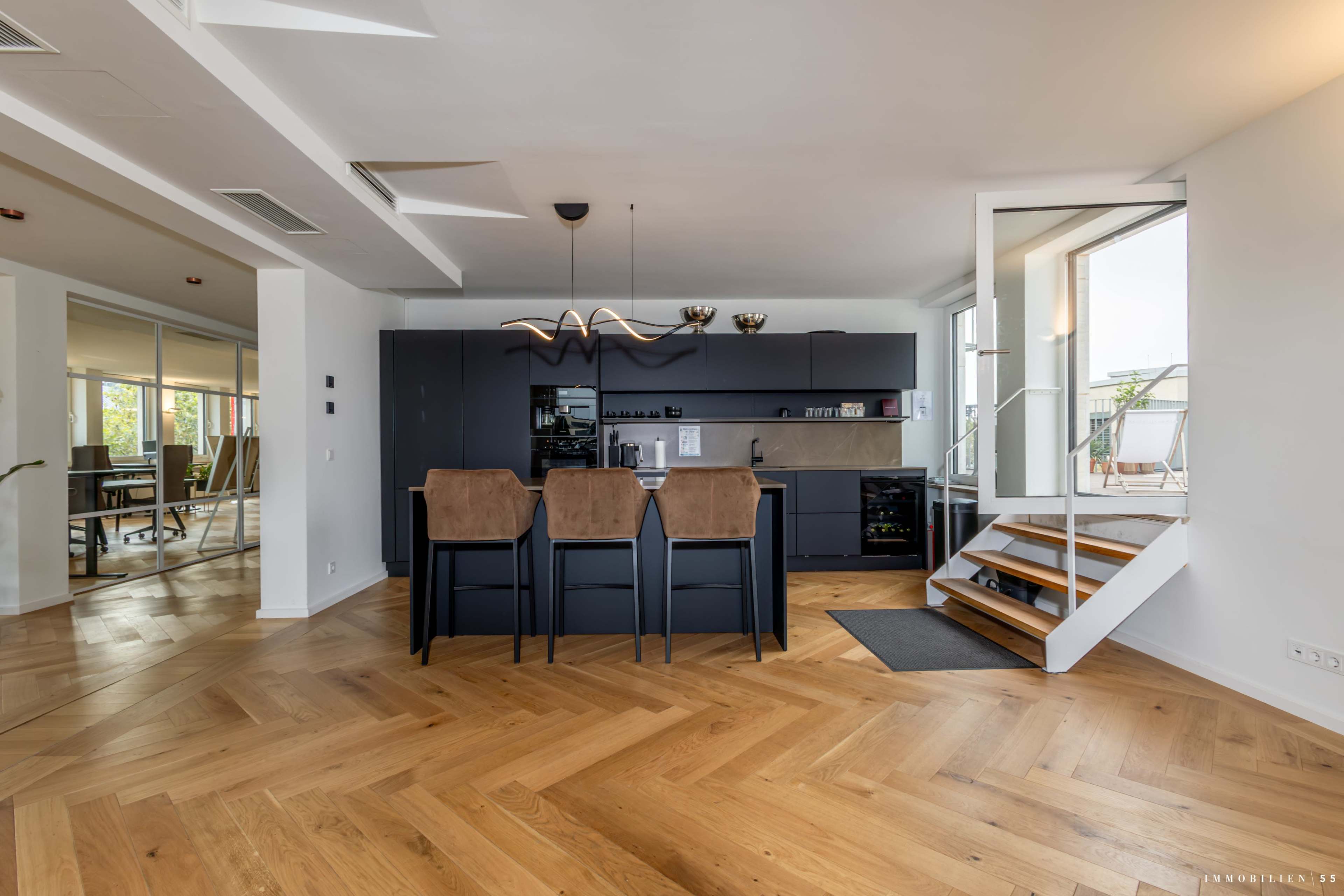 The image shows a modern kitchen area with dark cabinets, wooden flooring, and a staircase in a bright, open space.
