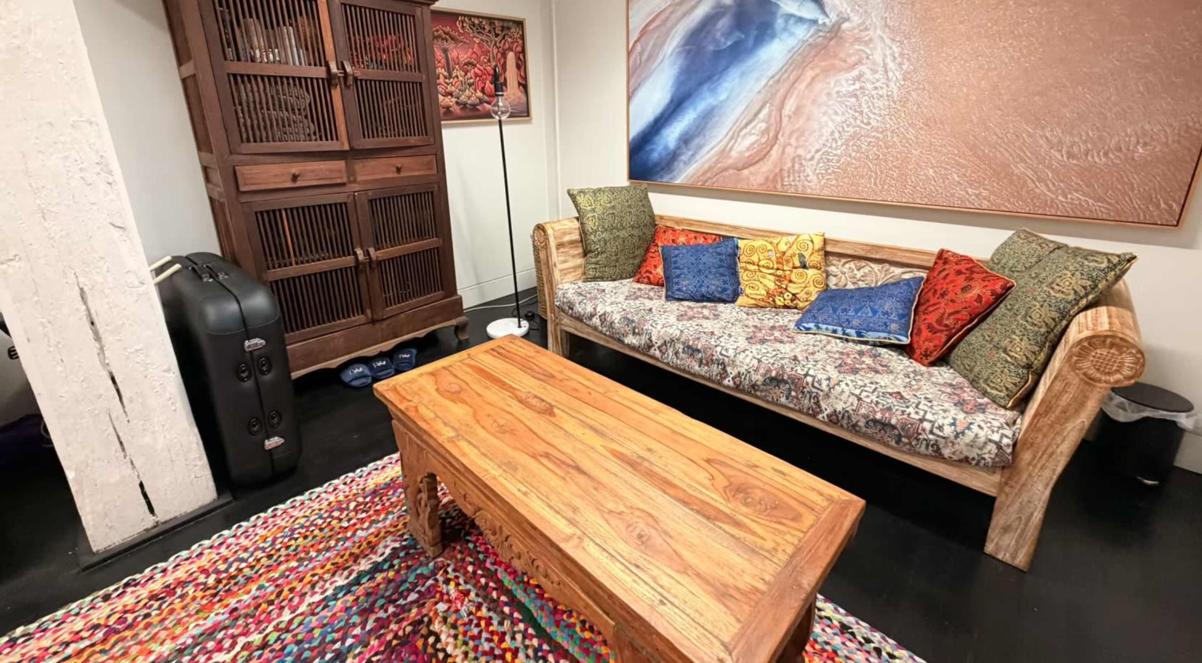 The image shows a cozy living room with a wooden sofa decorated with colorful cushions, a wooden coffee table, and a multicolored woven rug.