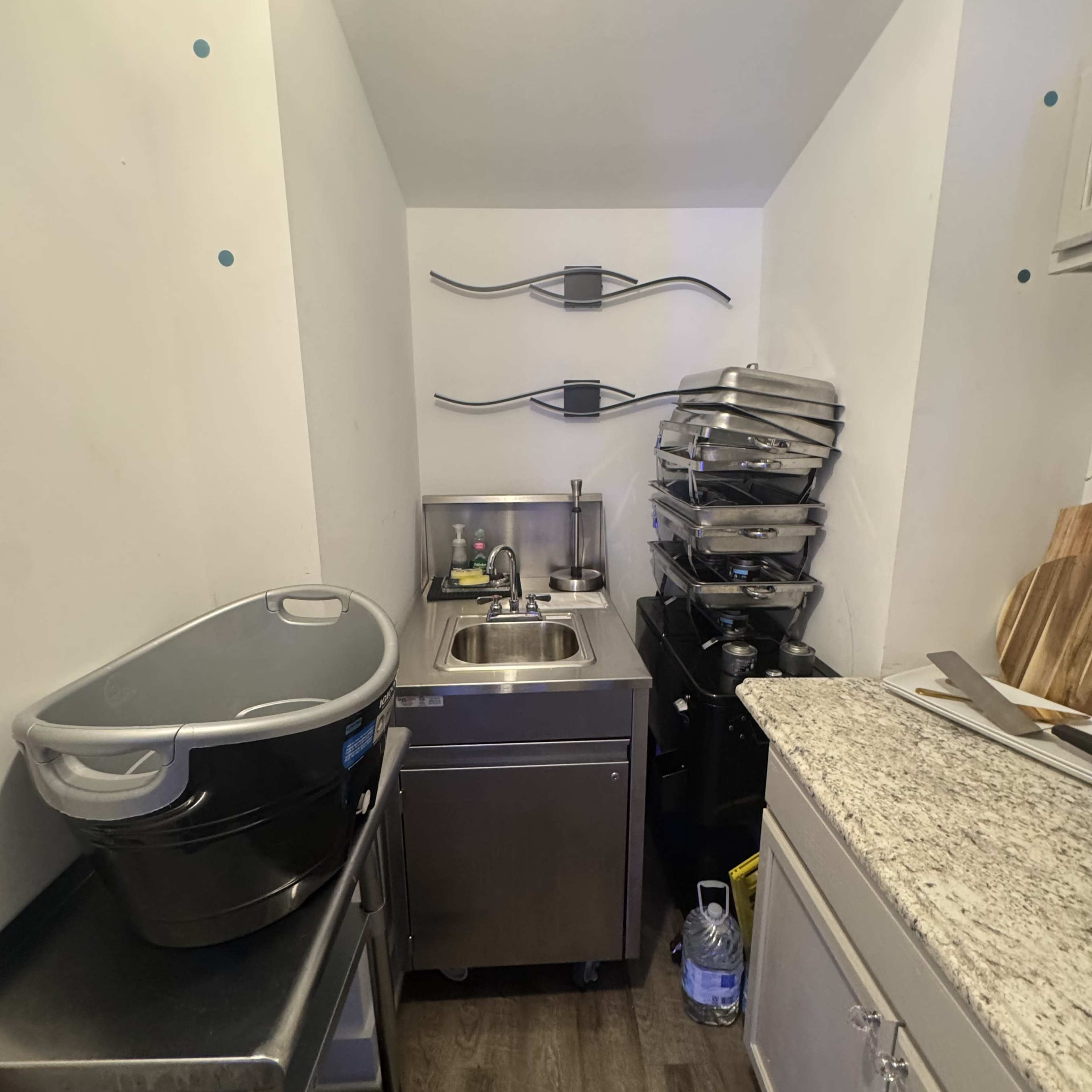 The image shows a small kitchenette with a sink, stacked food storage containers, and various kitchen tools on a countertop.