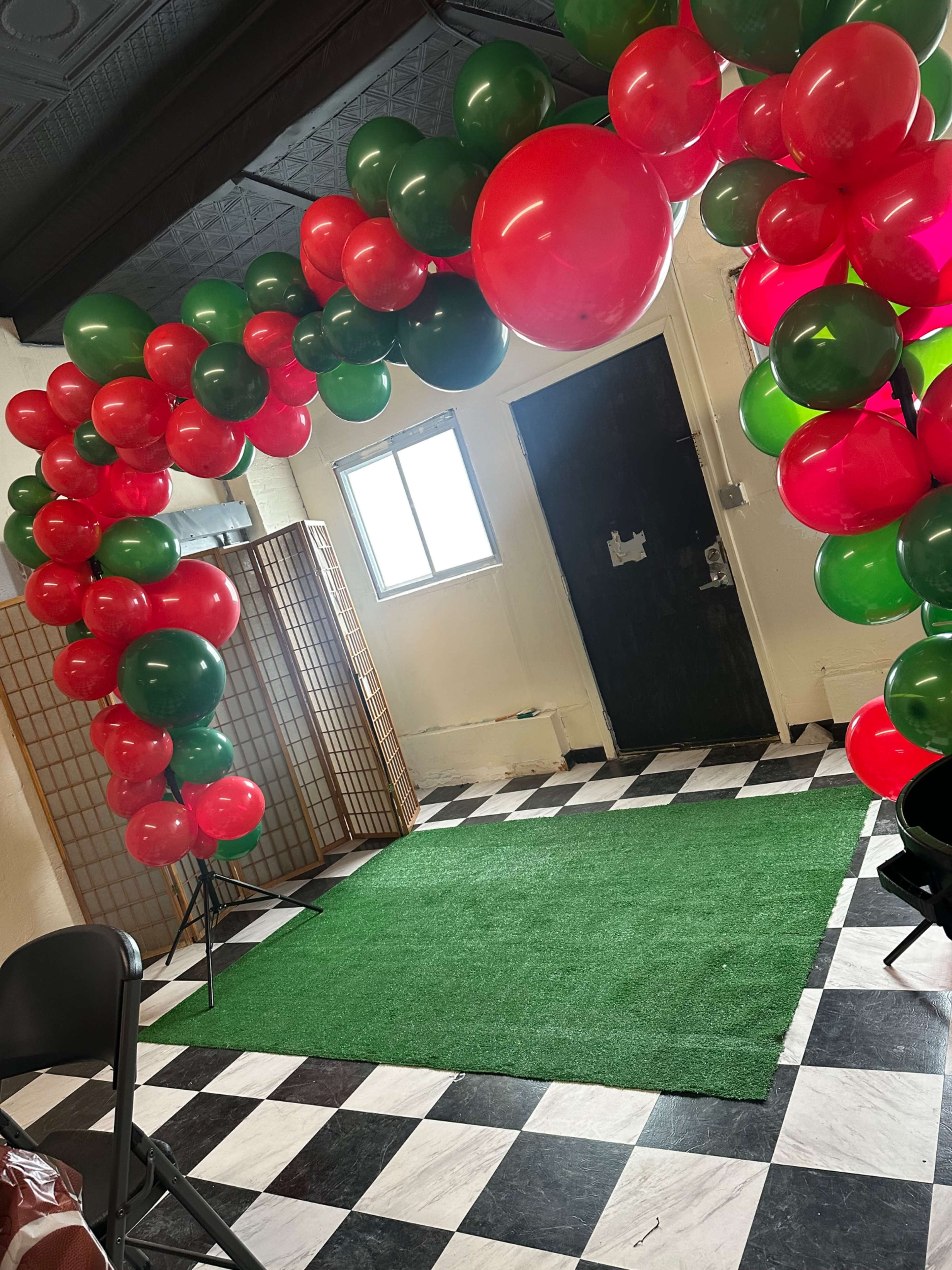 The image shows a decorated entrance arch made of red and green balloons, leading to an area with a green carpet and a checkered black and white floor.