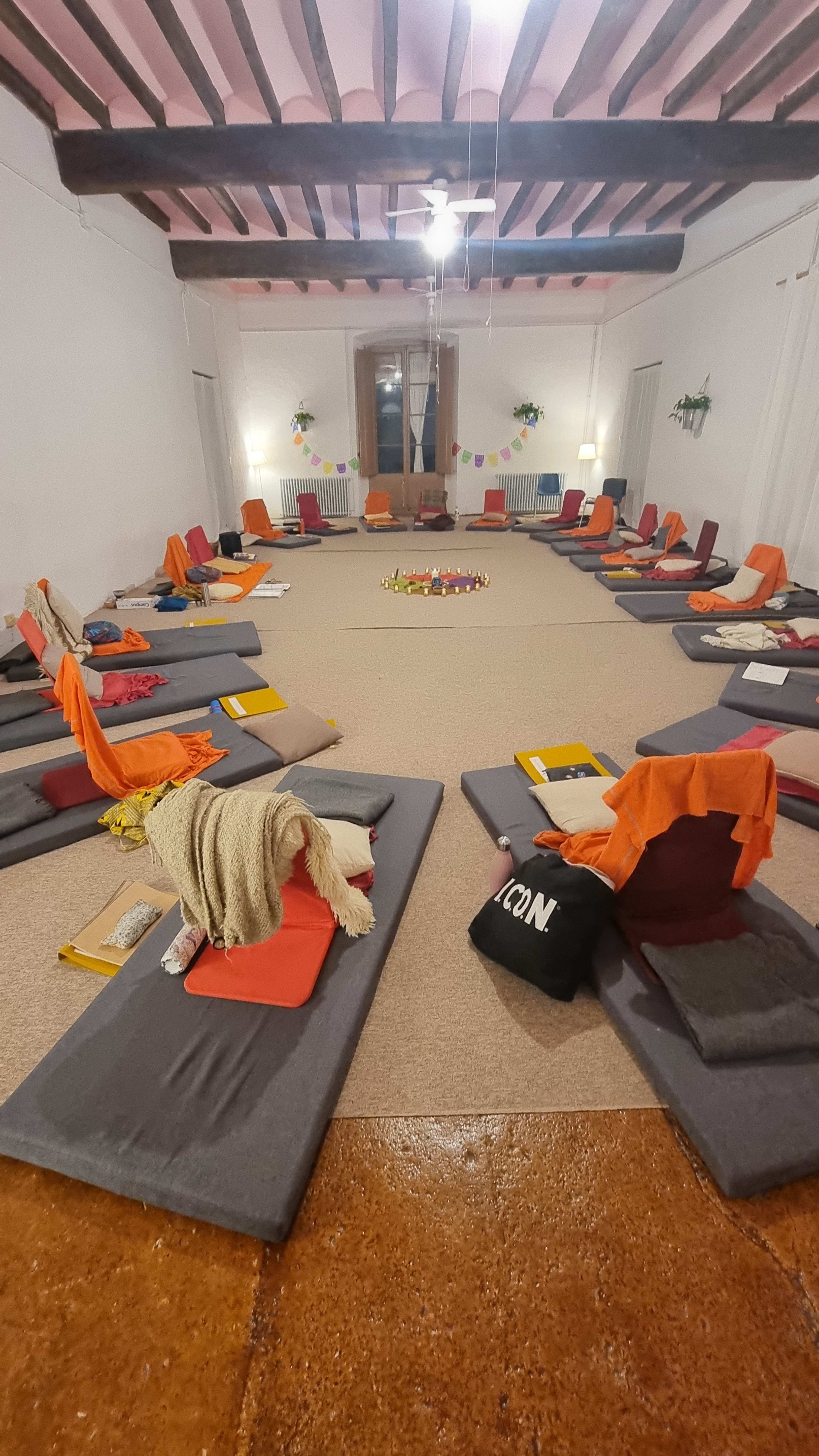 The image shows a room set up for a group activity, featuring arranged mats and cushions in a circular formation with candles in the center.