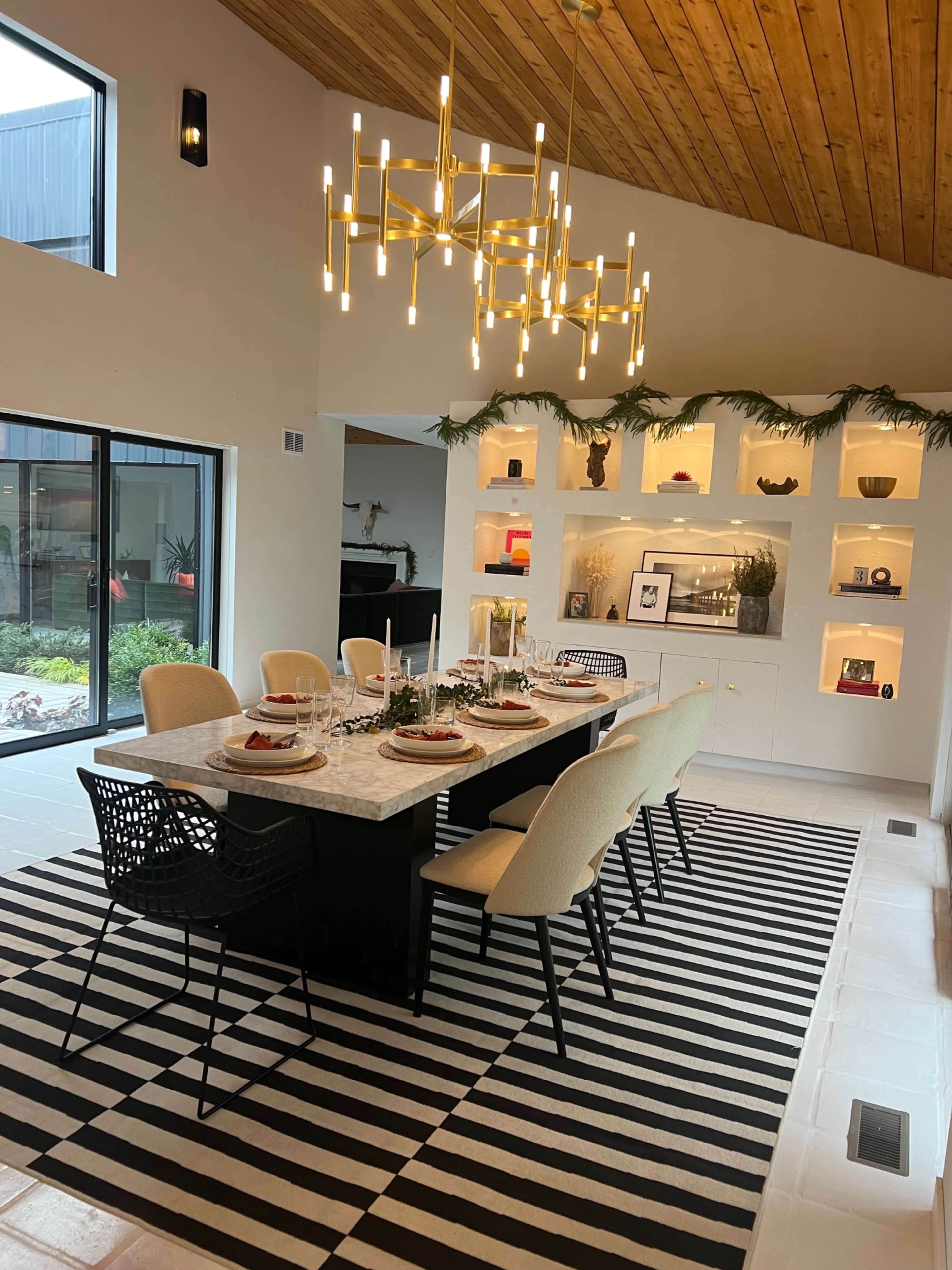 The image shows a modern dining room with a rectangular table set for a meal, surrounded by upholstered chairs, and accented by a stylish chandelier and decorative shelving in the background.