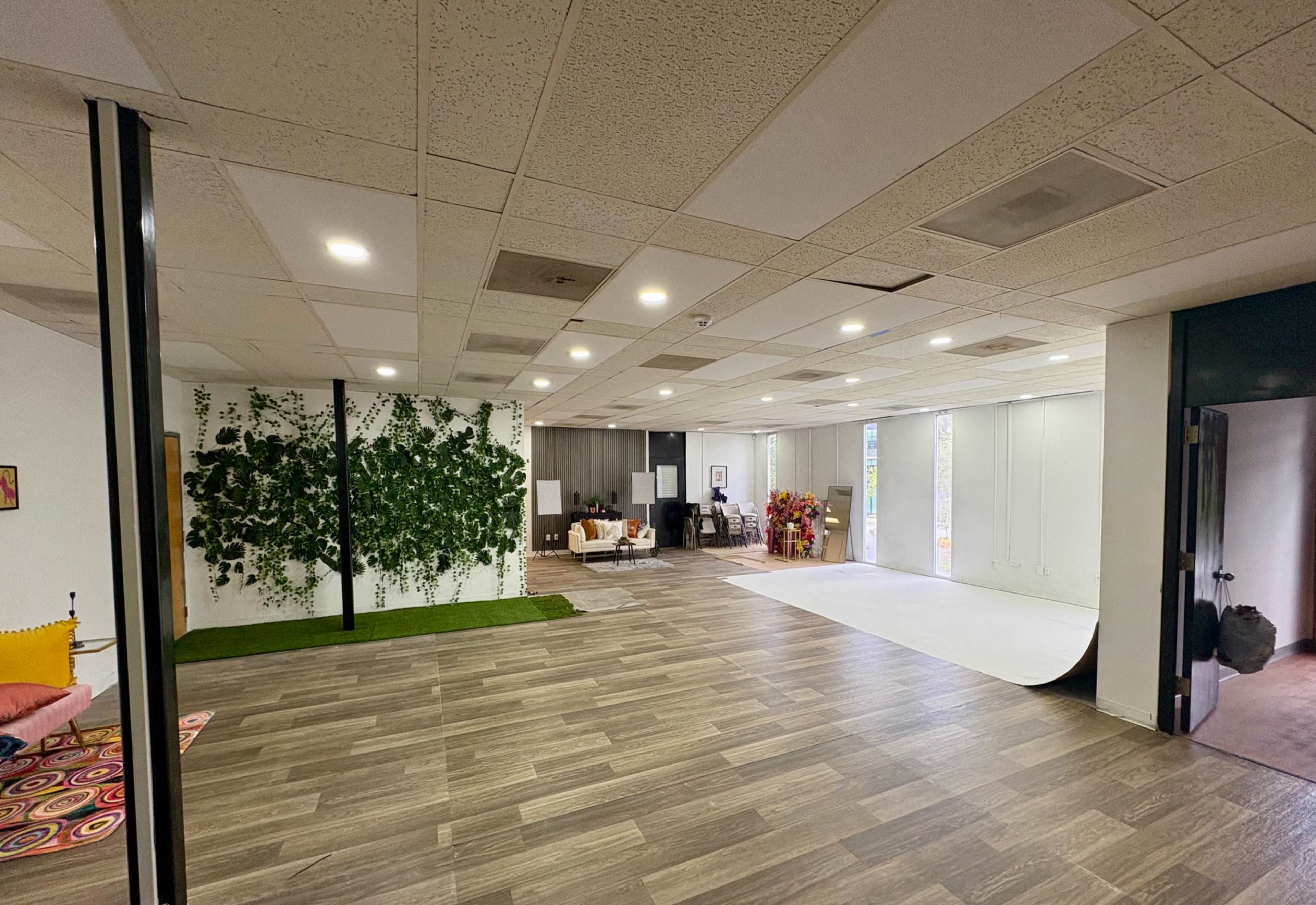The image shows a spacious, modern office area featuring a green wall of plants, wooden flooring, and various furniture arrangements in distinct sections.