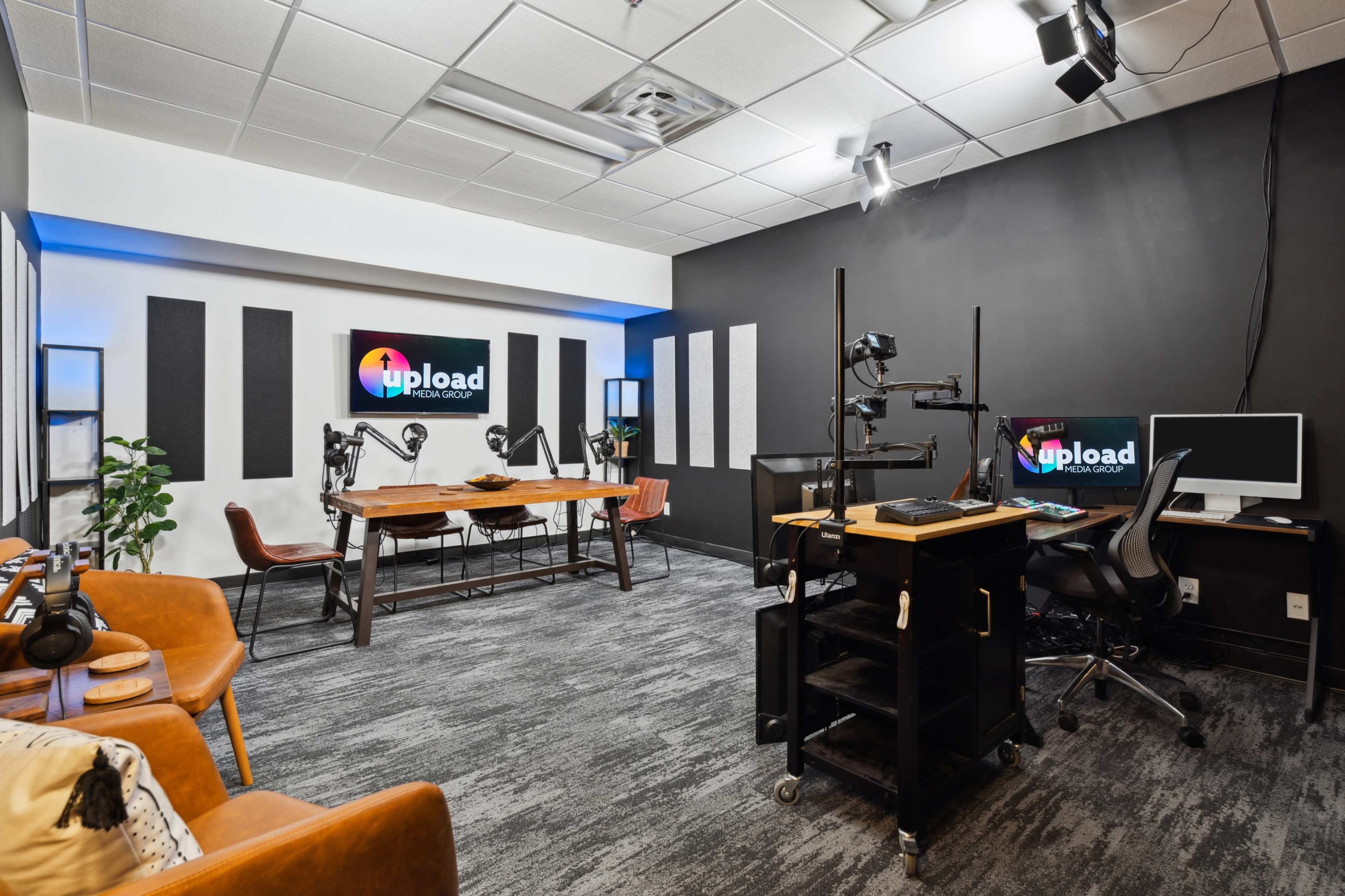 The image shows a modern studio space featuring a large wooden table, multiple camera setups, a computer workstation, and dark walls with sound panels.