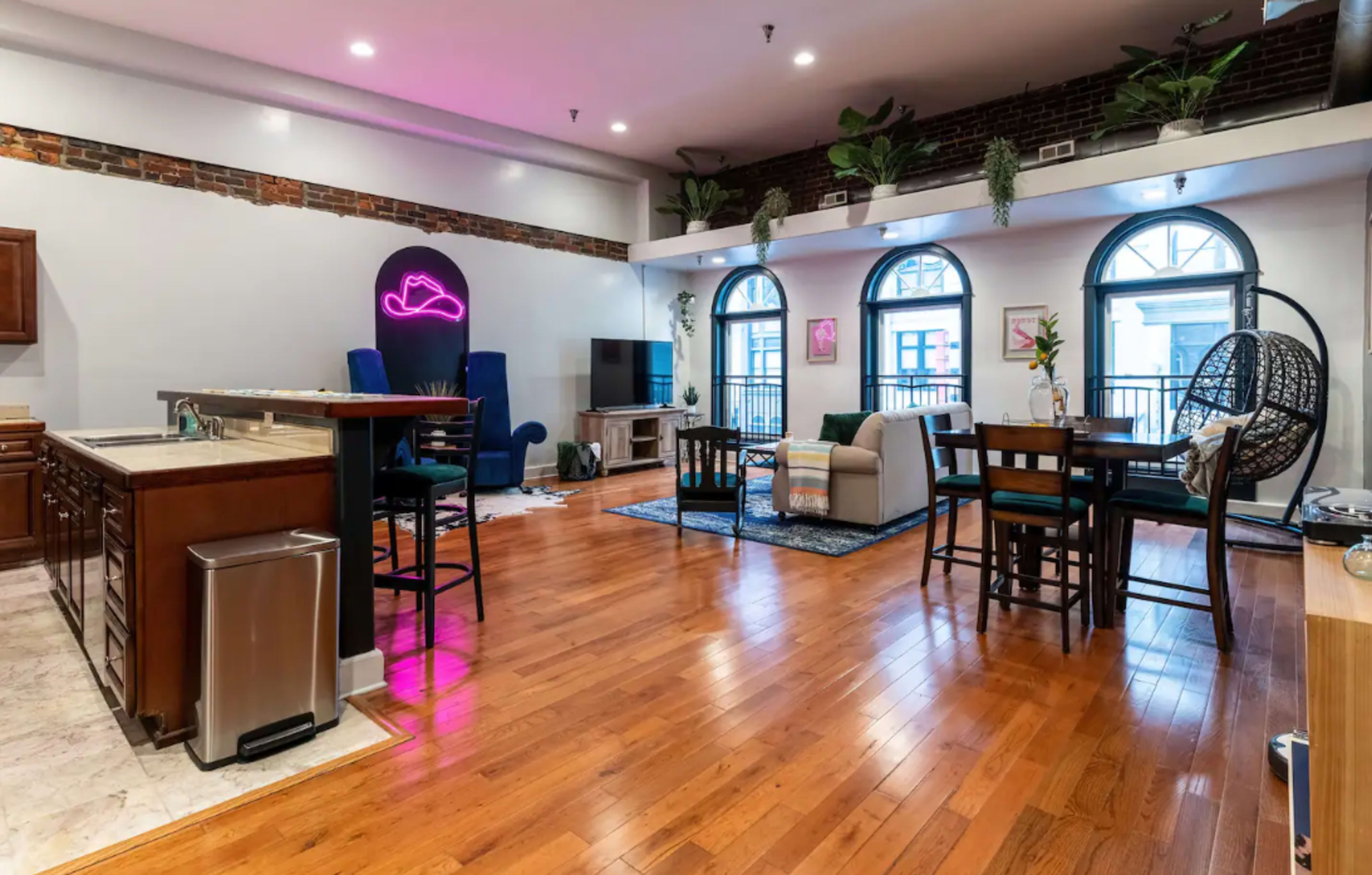 The image shows a spacious living area with hardwood floors, a kitchen island, seating arrangements, and large windows illuminated by indoor plants and ambient lighting.