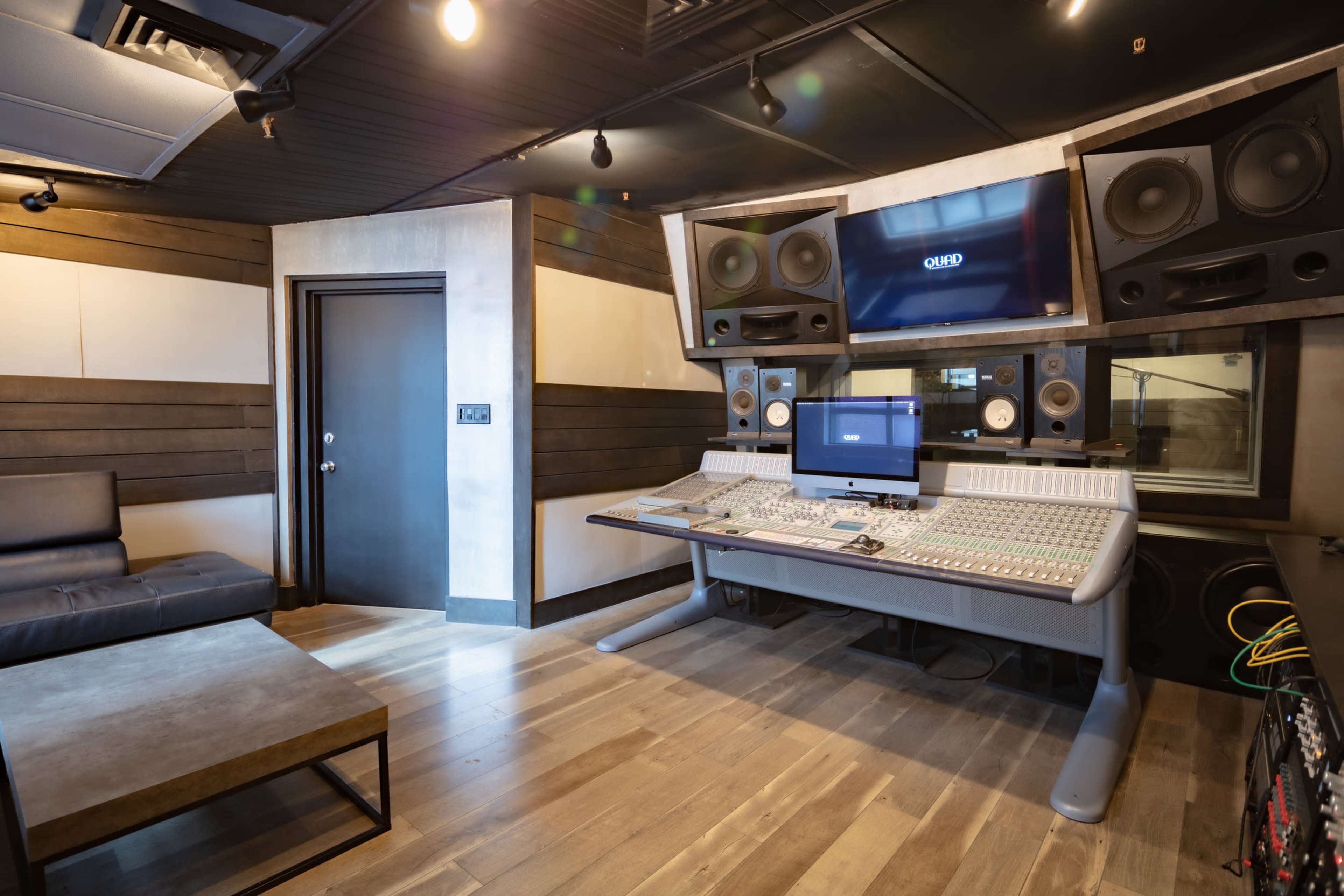 The image shows a spacious music recording studio equipped with a large mixing console, monitors, and a seating area.