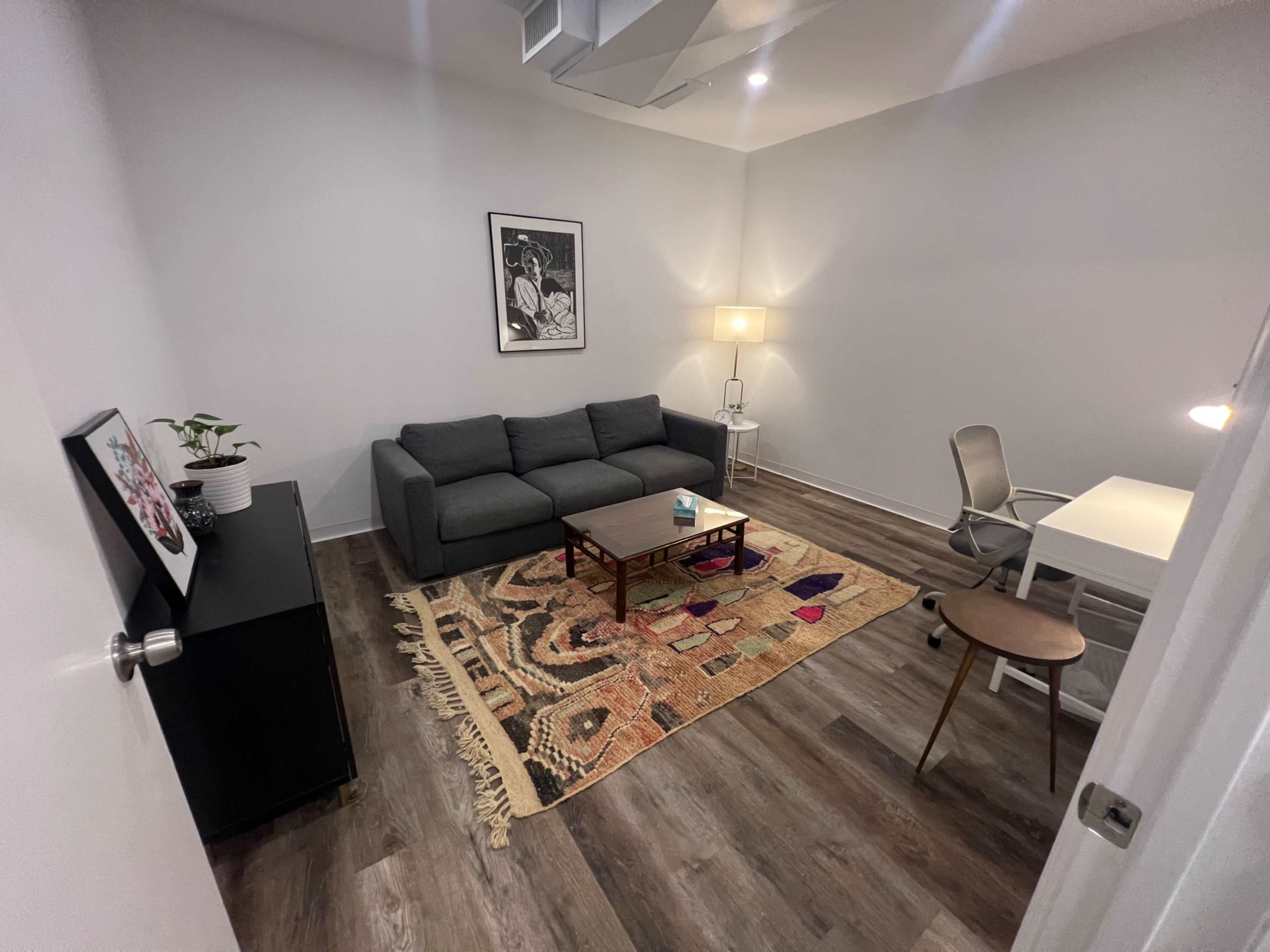 A small, modern room features a gray sofa, a coffee table on a patterned rug, a desk with a chair, and a lamp.