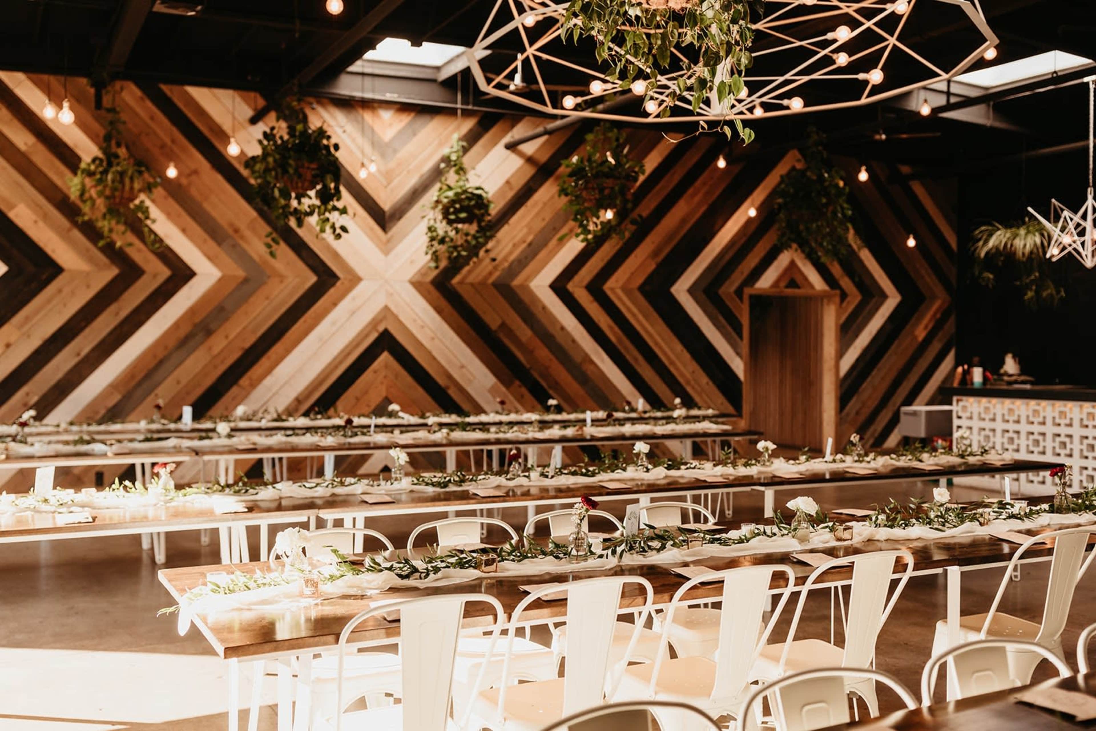 The image shows a modern event space with long tables decorated with greenery and flowers, set against a striking chevron-patterned wooden wall.