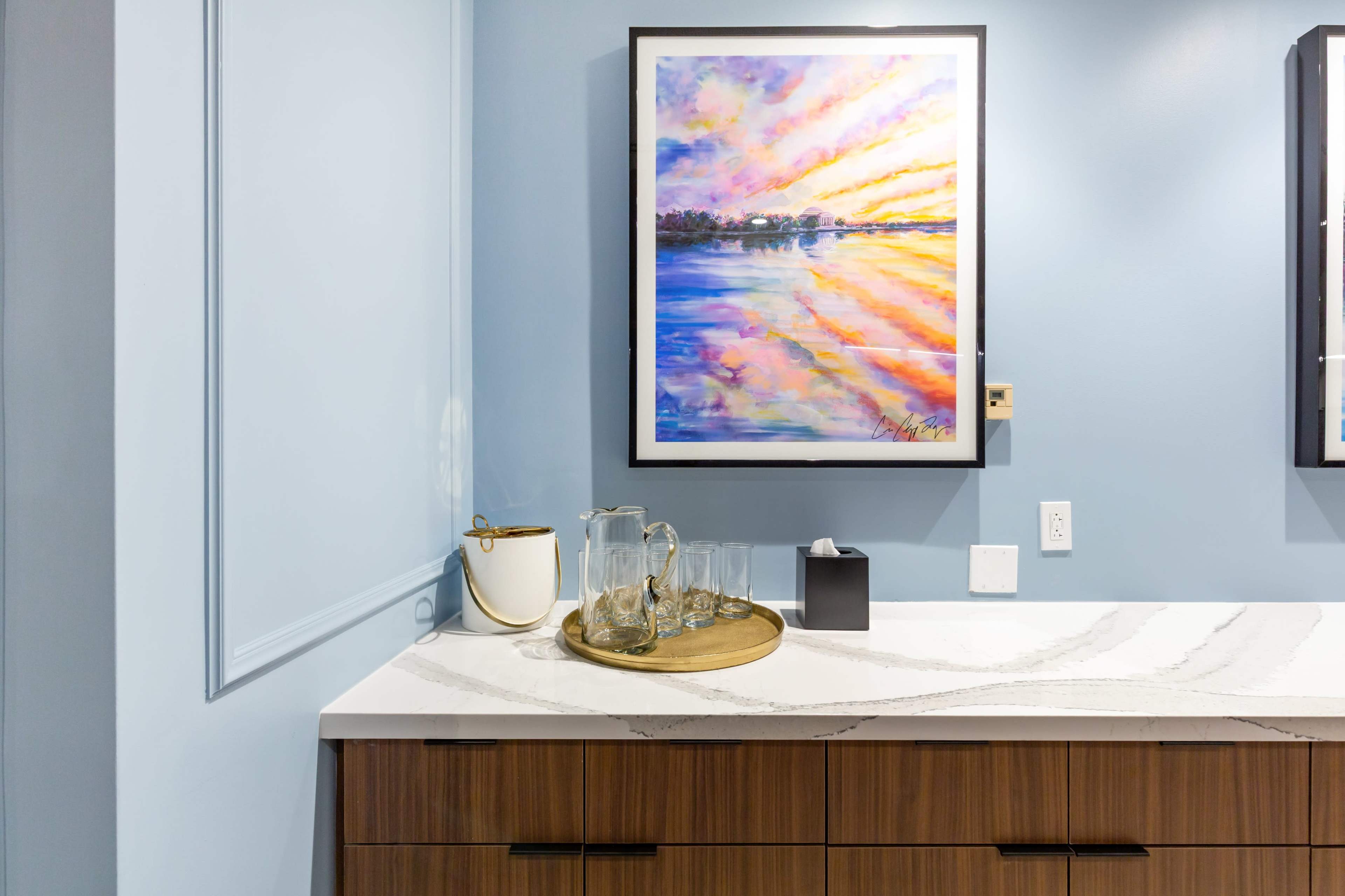 The image shows a modern countertop with a tray containing glassware, a napkin holder, and a framed colorful painting hanging on the wall above a light blue backdrop.