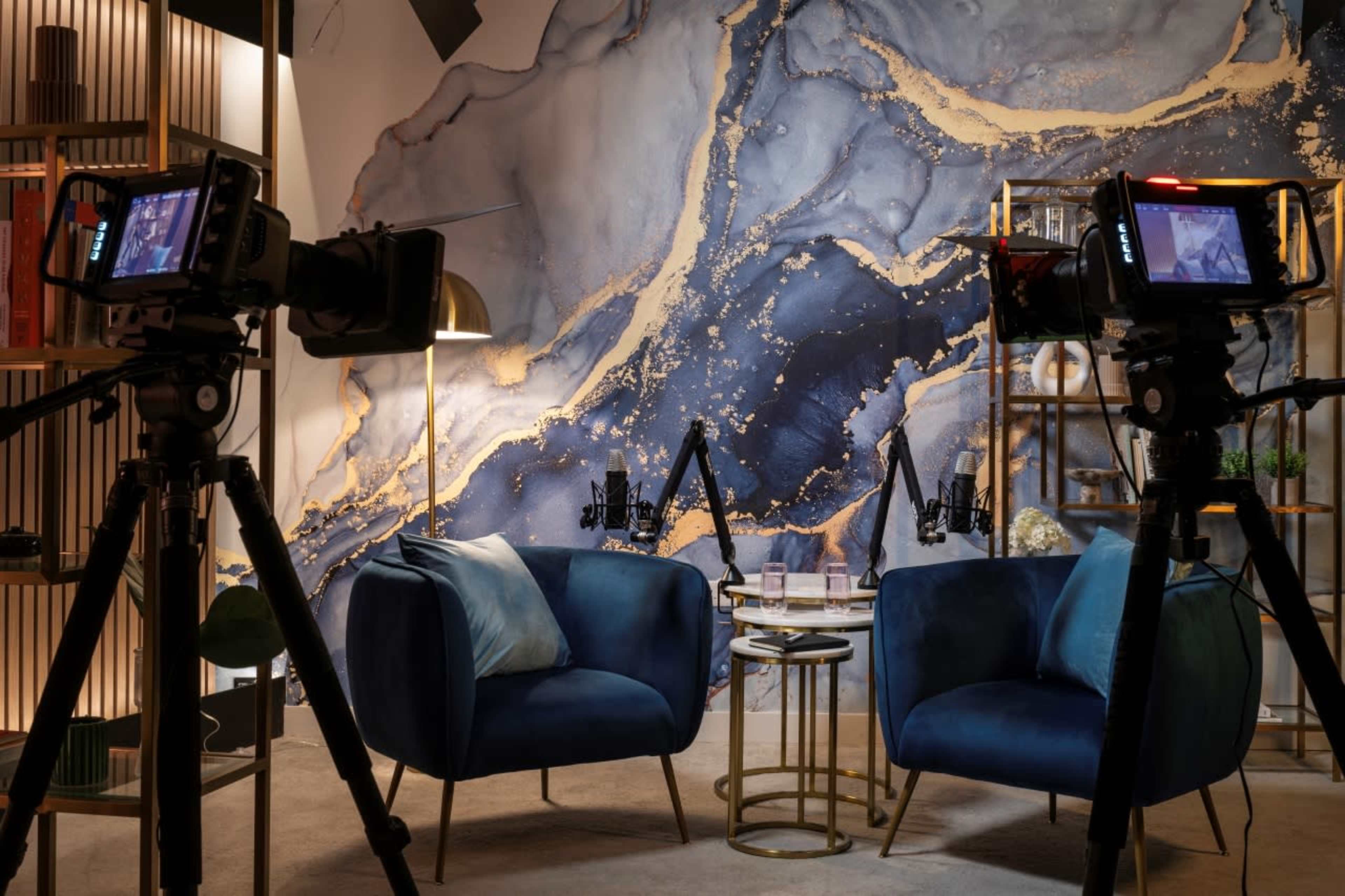 A modern podcast studio with two blue chairs facing a small table, surrounded by cameras and a decorative wall featuring abstract patterns.
