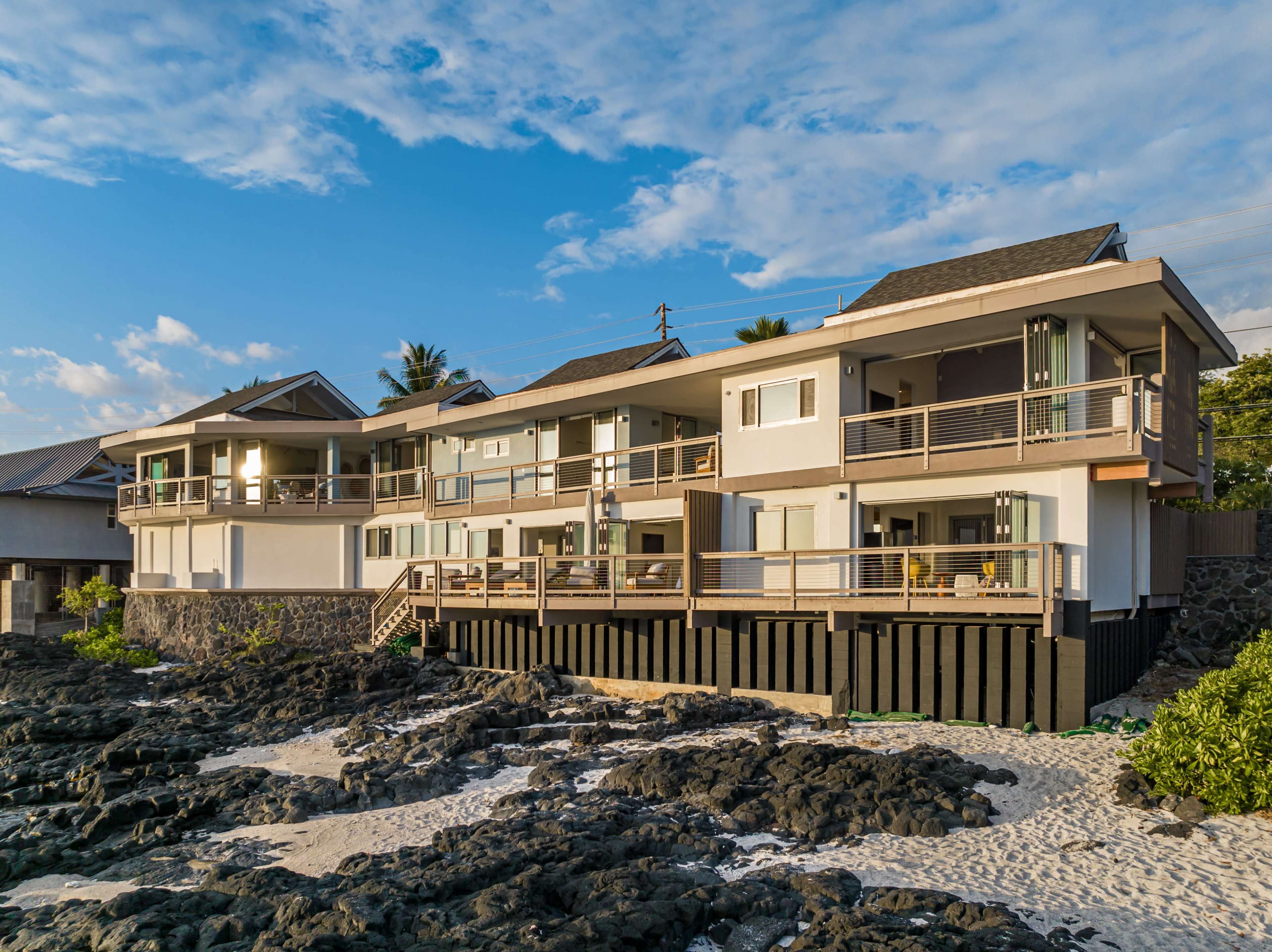A modern beachfront house with multiple levels stands on a rocky shoreline, surrounded by sand and under a clear blue sky.