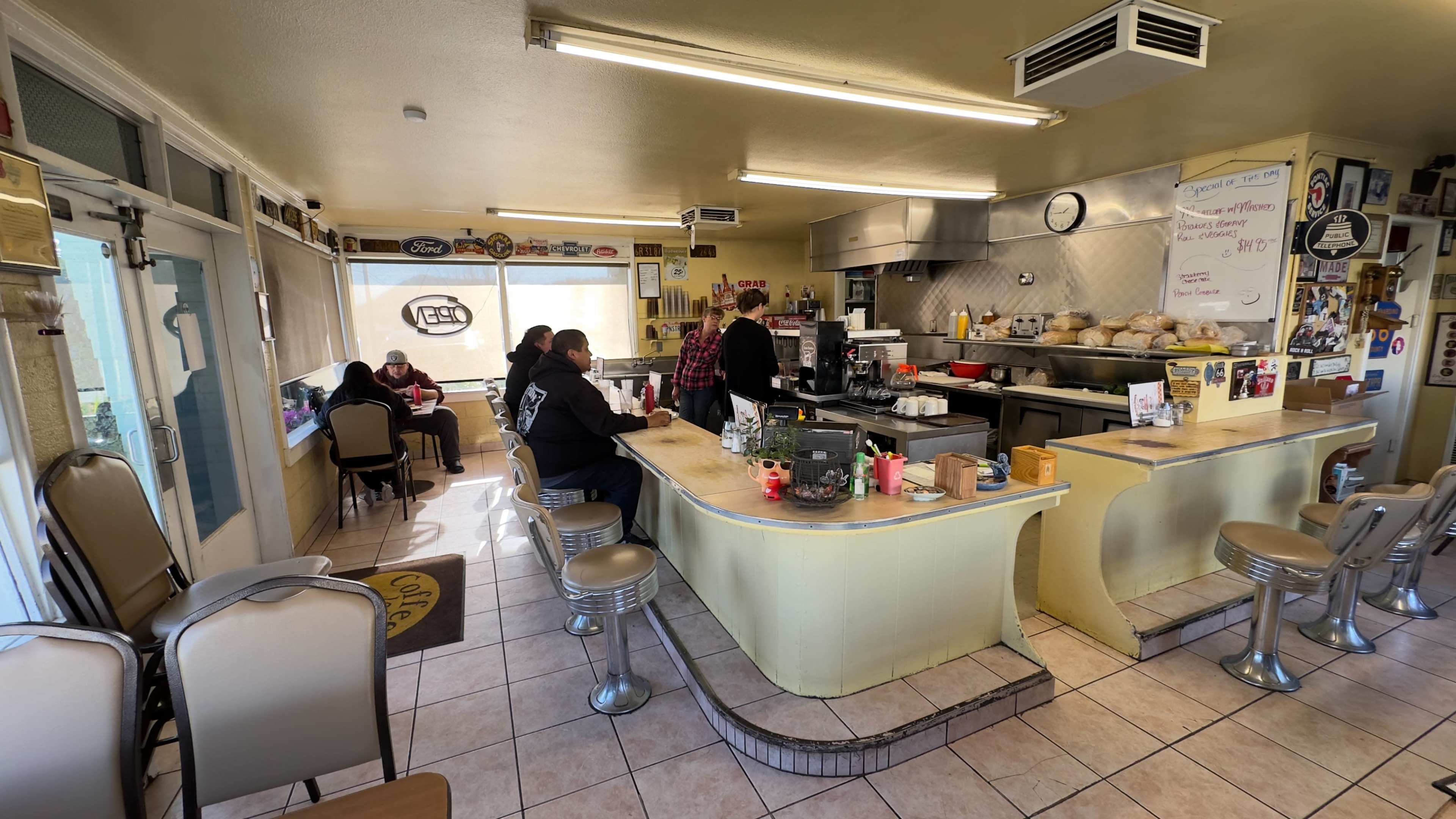 A small diner features a counter with stools, a grill area, and several patrons seated at tables.