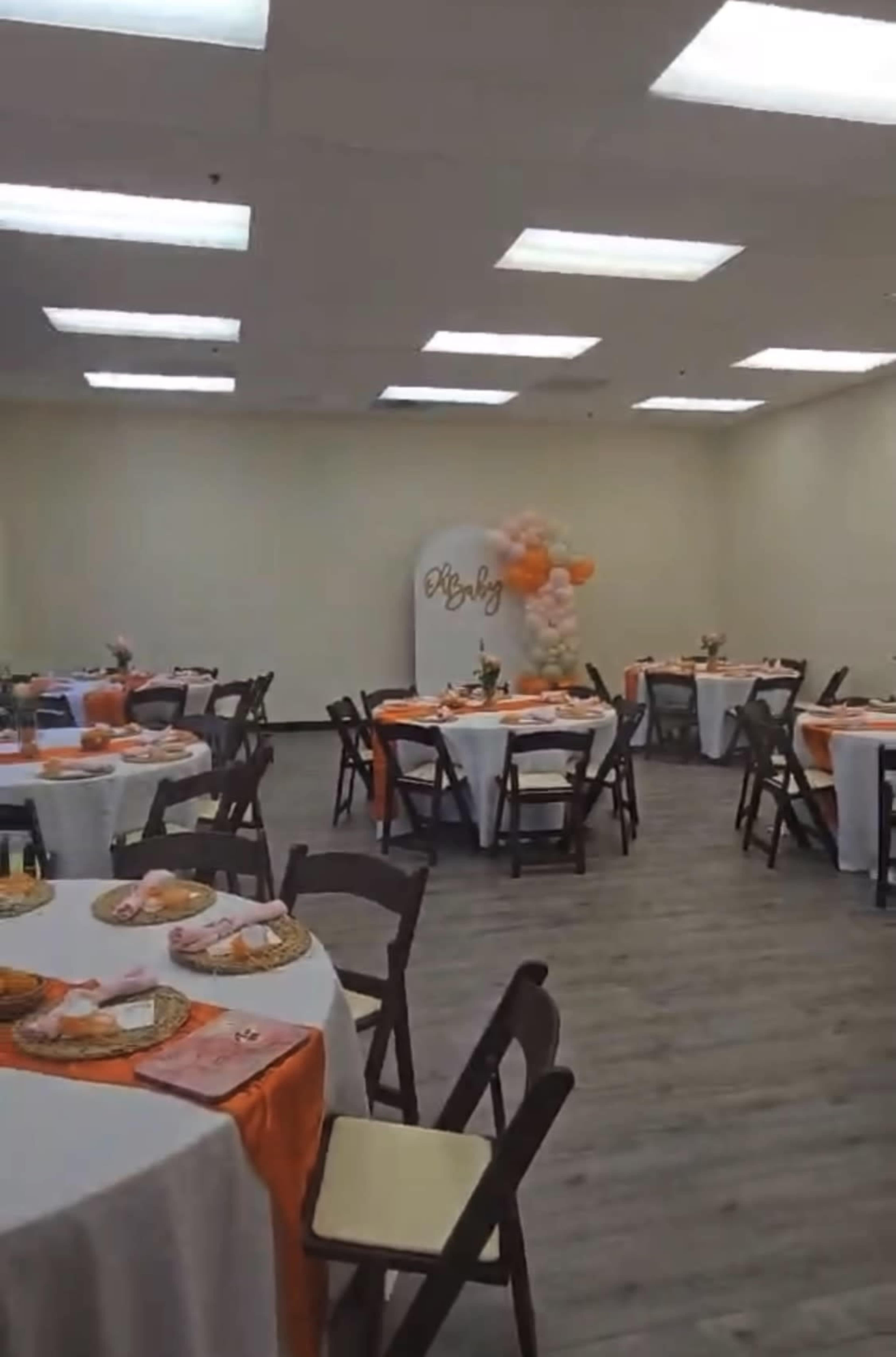 The image shows a decorated event space with multiple round tables set with orange and white tablecloths, featuring floral centerpieces and plates of food.
