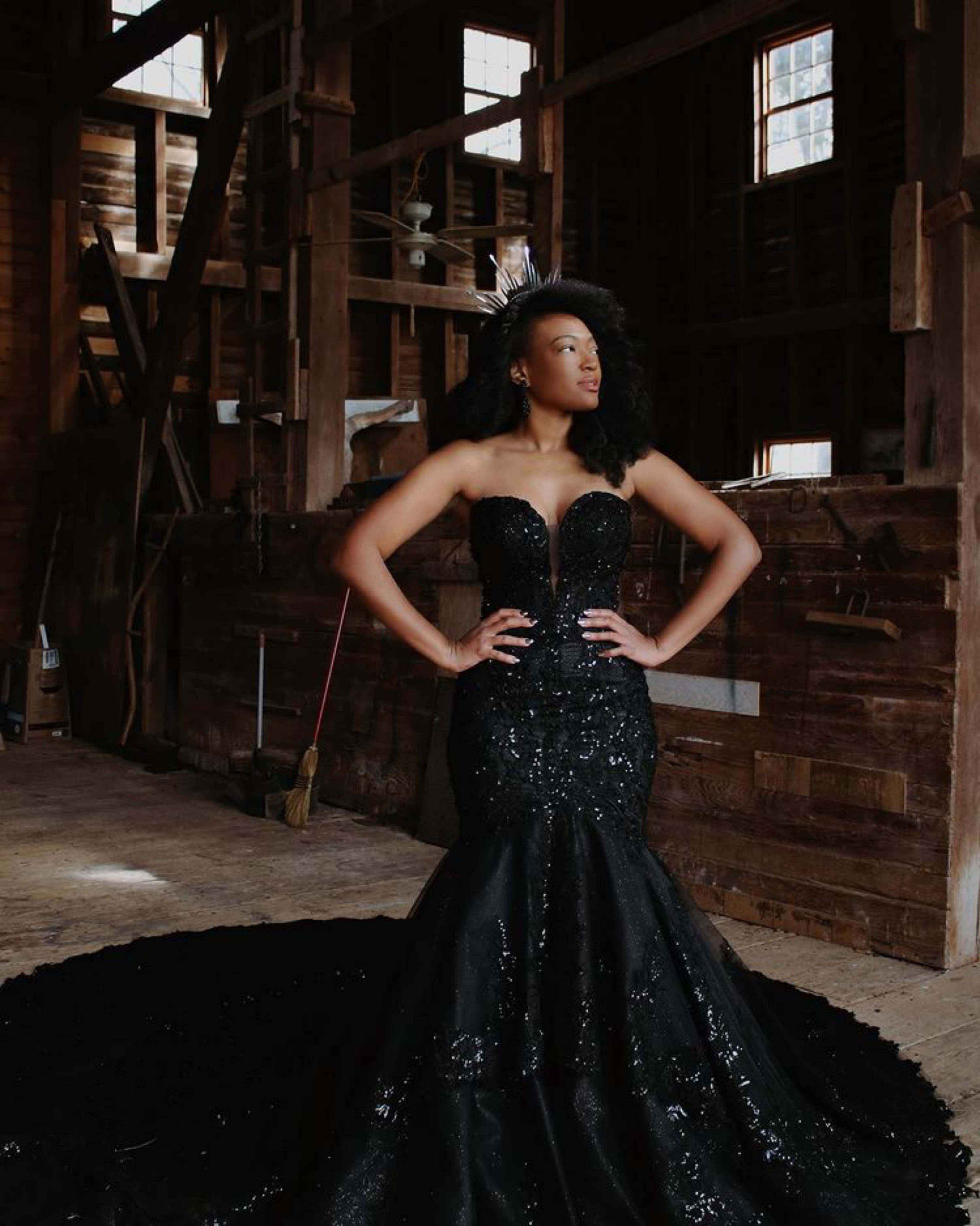 A woman in a form-fitting black gown with a train poses confidently in a rustic wooden barn setting.
