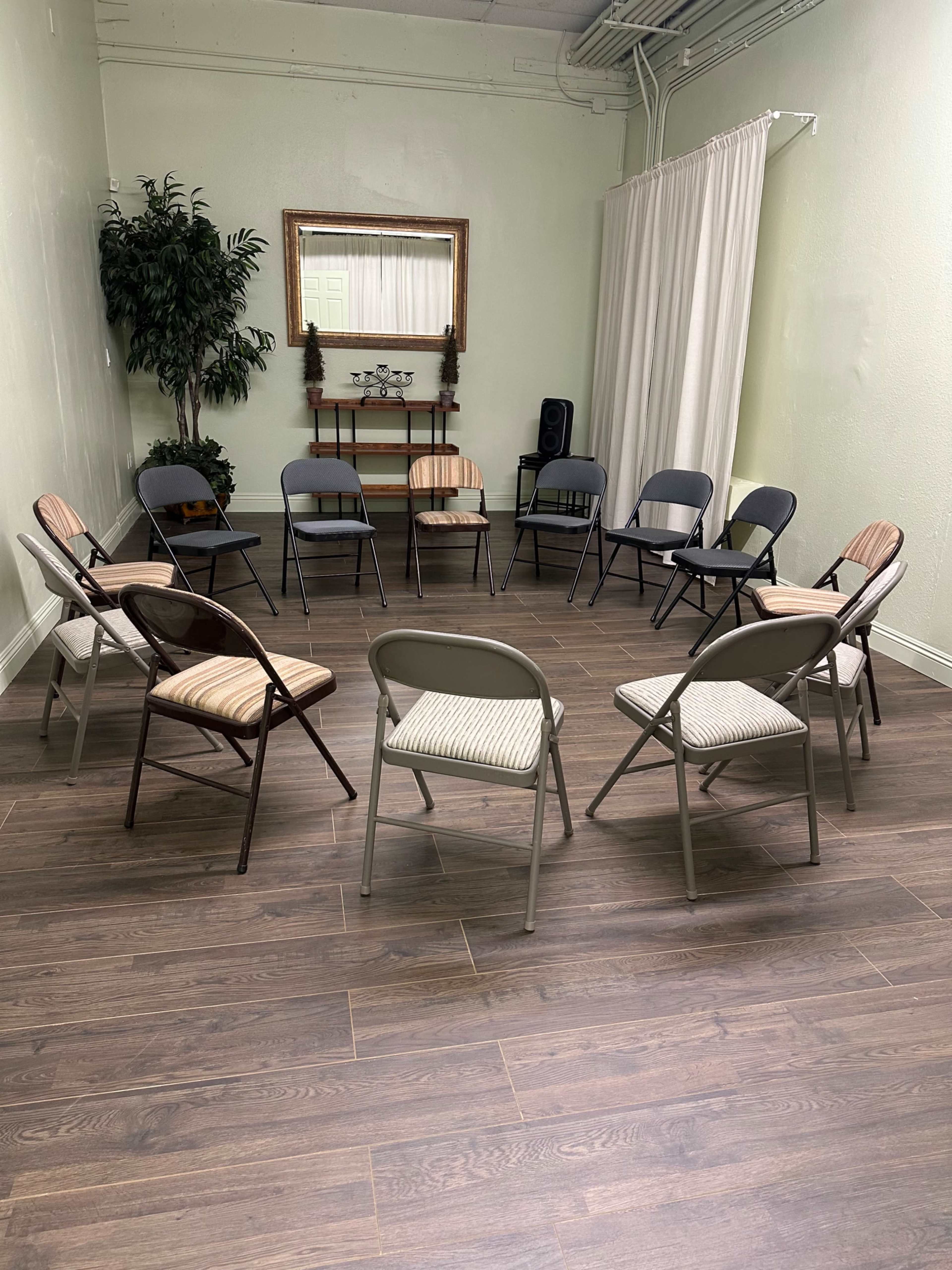 The image shows a circular arrangement of various folding chairs in a minimalistic room with a mirror, a plant, and a shelf in the background.