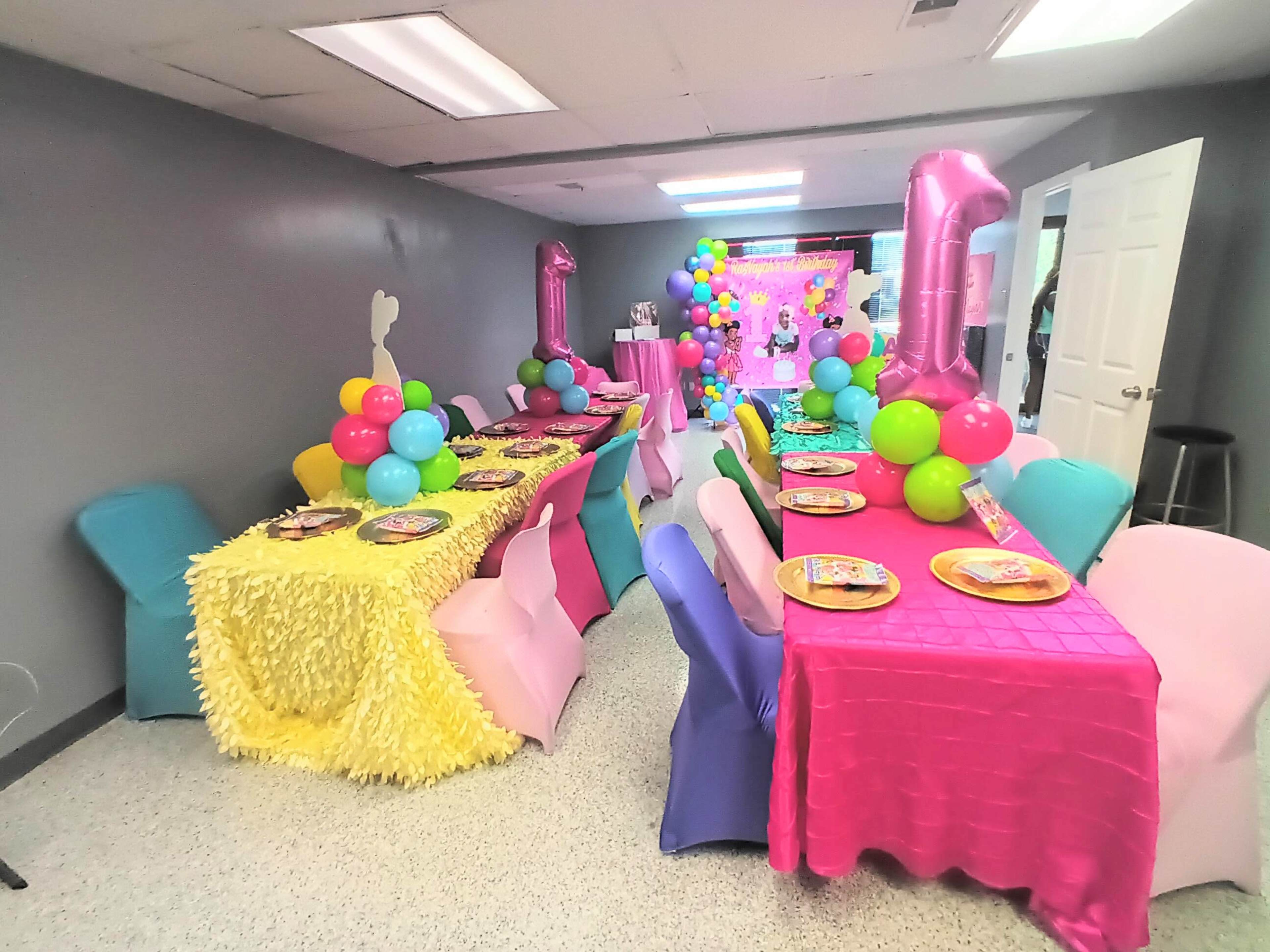 A festive party room features tables covered in colorful tablecloths adorned with balloons and themed decorations.