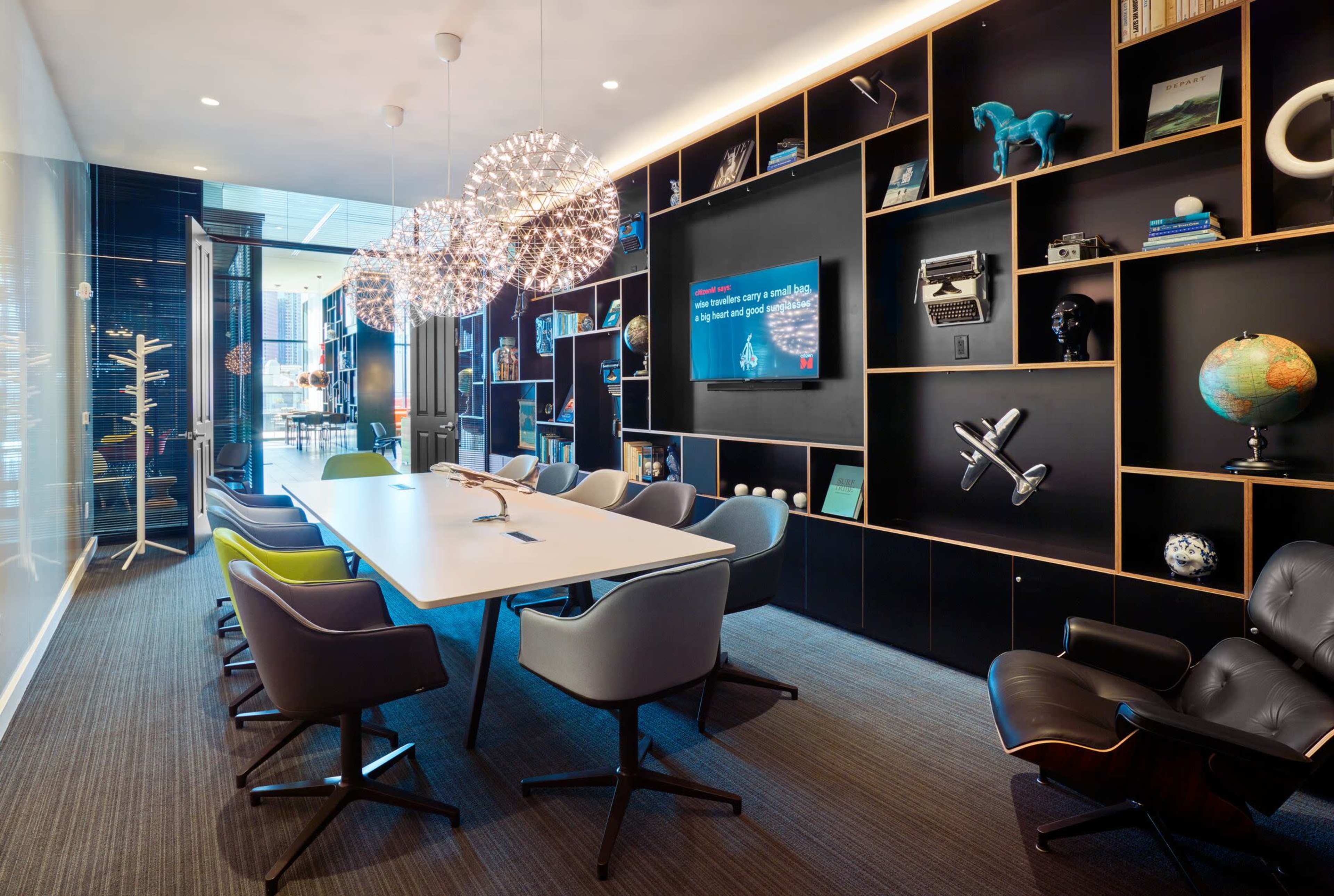 A modern conference room features a long white table surrounded by stylish chairs, illuminated by hanging light fixtures, with a black shelving unit displaying decorative items and technology.