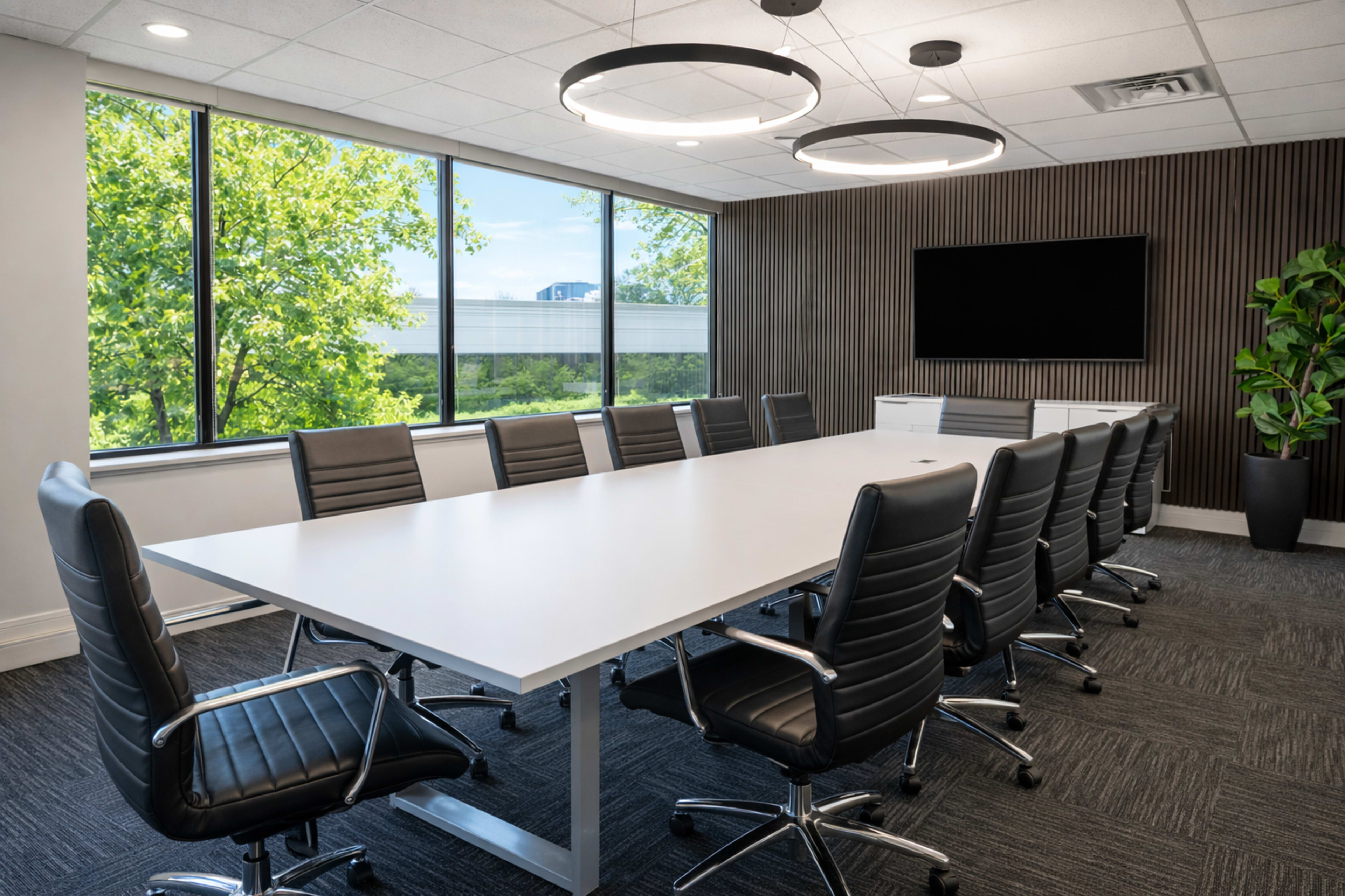 A modern conference room features a long white table surrounded by black leather chairs, large windows offering a view of trees, and a wall-mounted television.