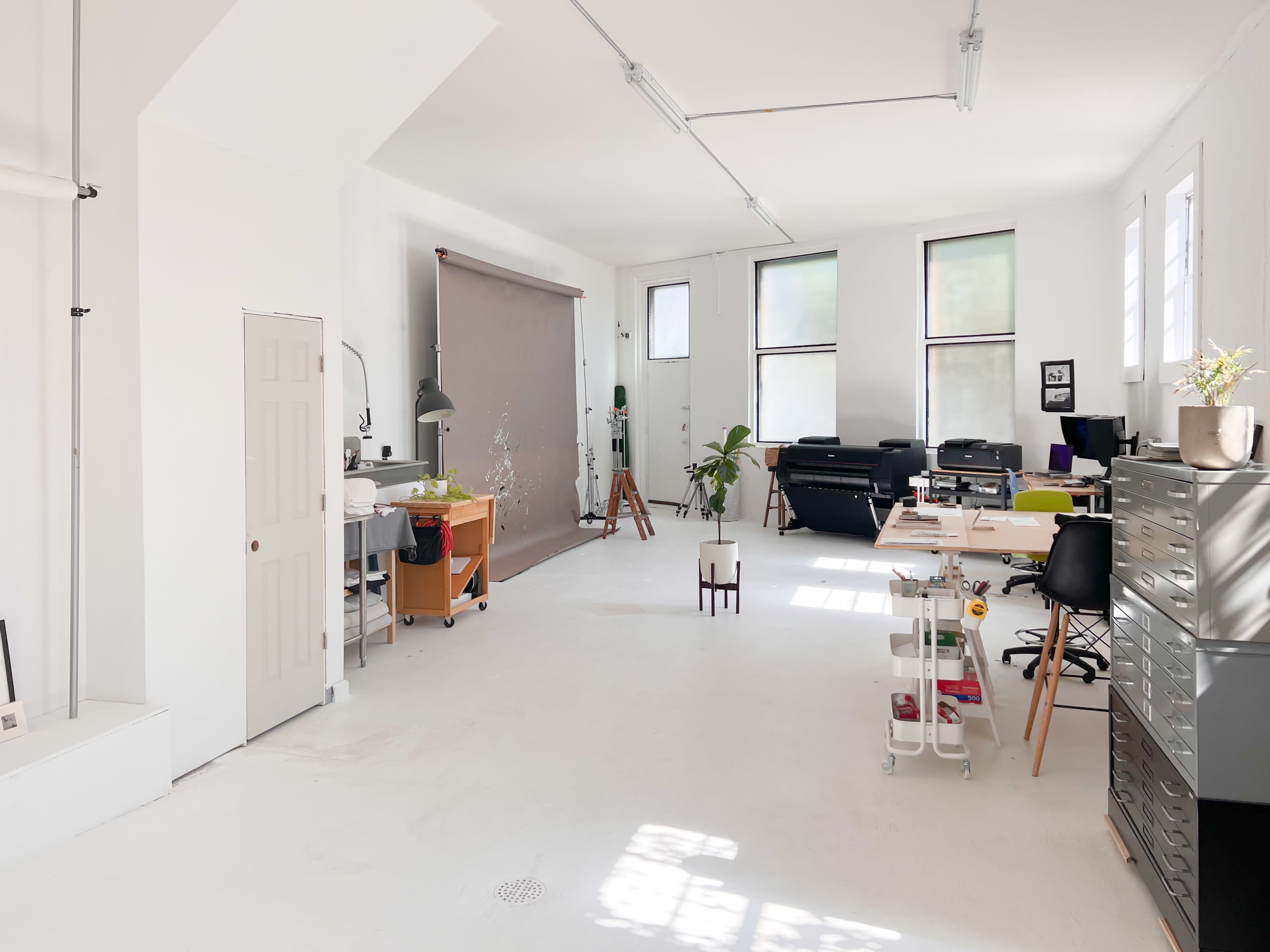 A spacious, well-lit studio featuring white walls, a large backdrop, and various workstations with furniture and equipment.