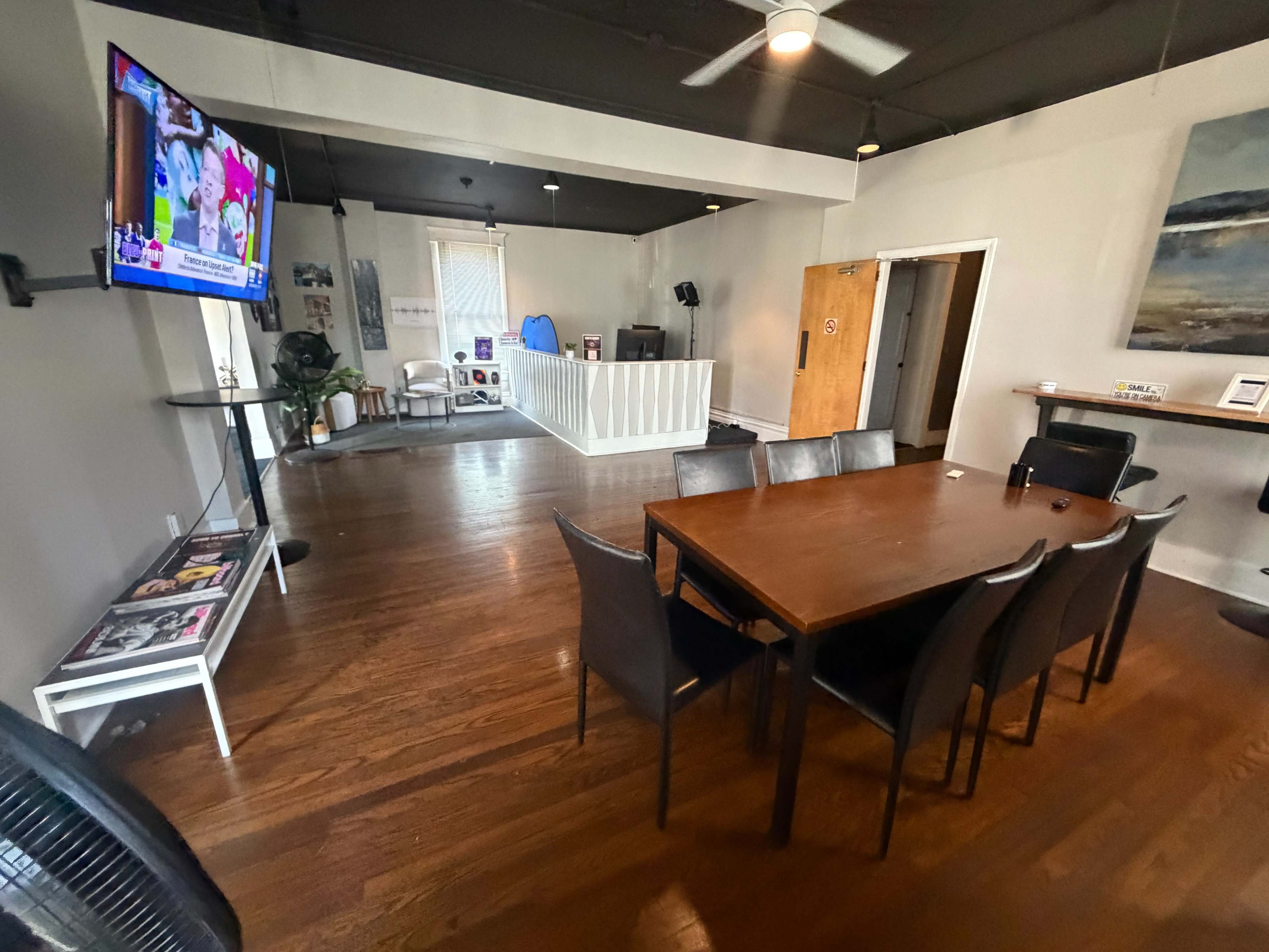The image shows a spacious room with a wooden table surrounded by black chairs, a TV mounted on the wall, and a few seating areas along with a white partition in the background.
