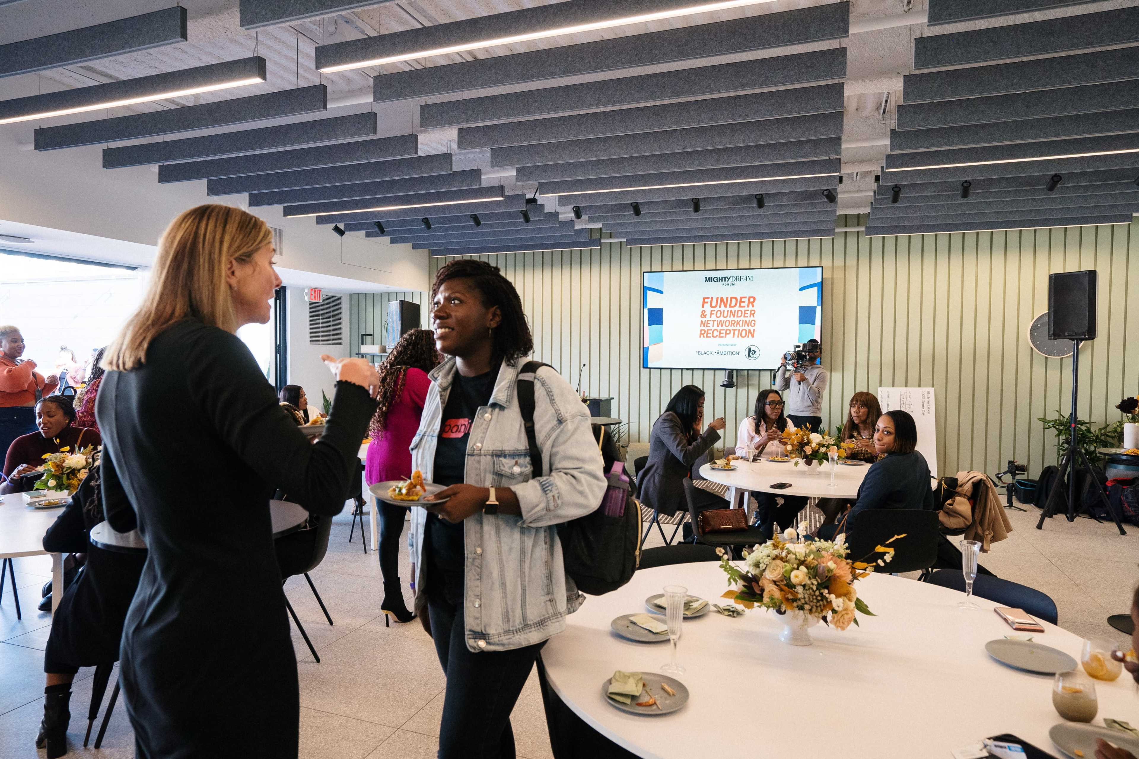 Two women are engaged in conversation at a networking event featuring tables with food and floral arrangements.