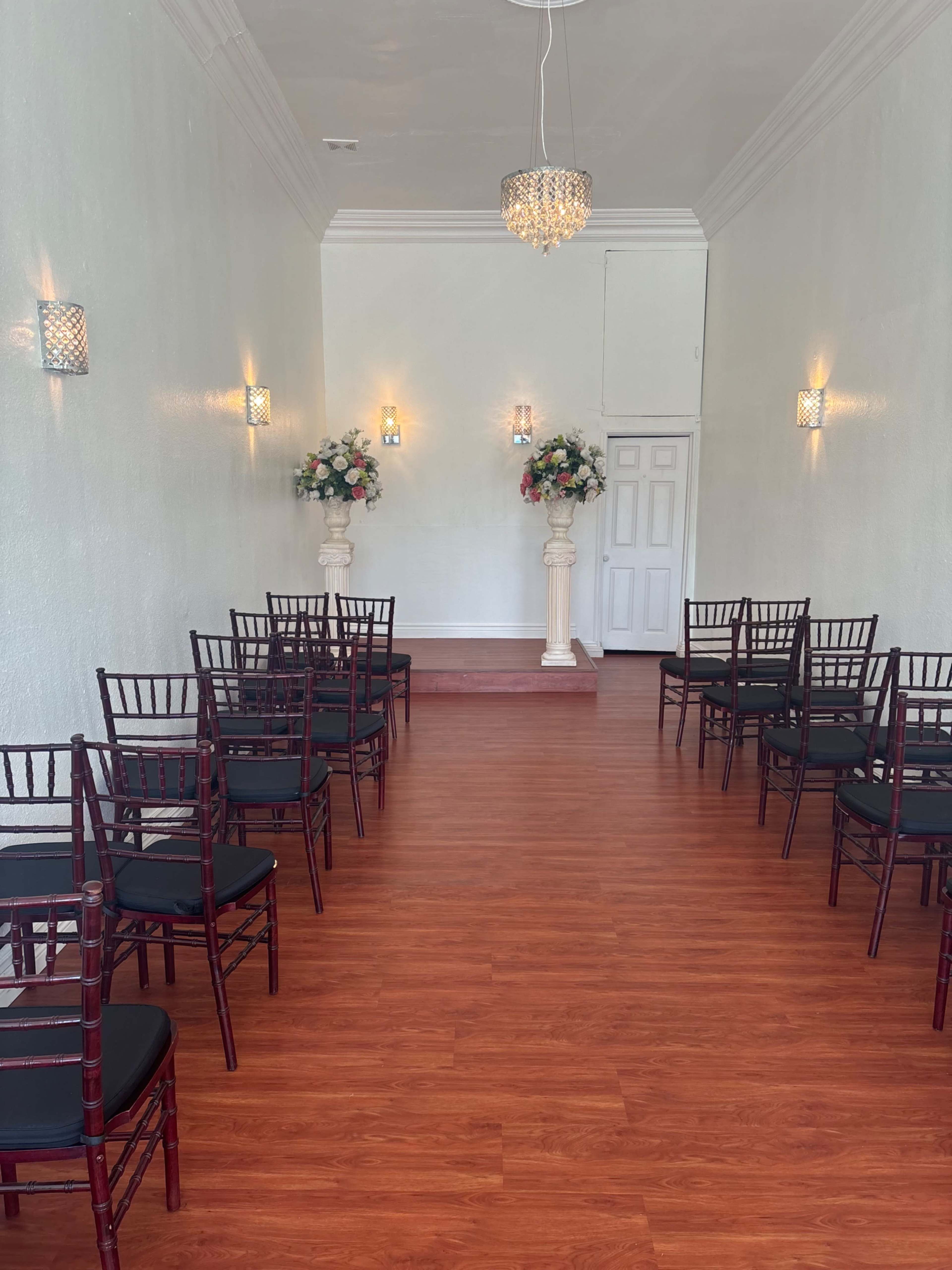 The image shows a narrow, well-lit room set up for a formal event, featuring rows of chairs facing a small stage with floral arrangements on pedestals.