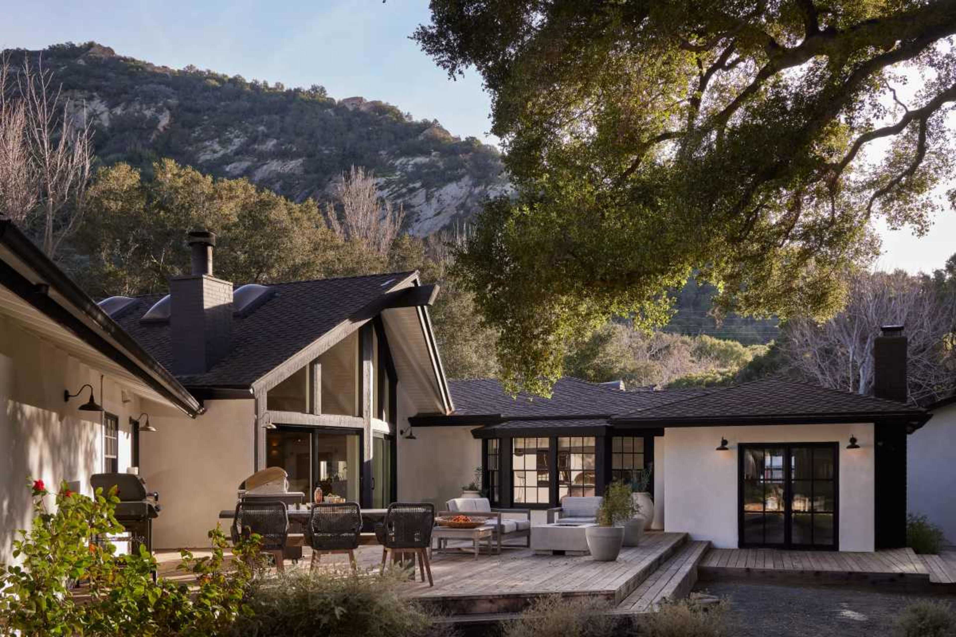 A modern home with a wooden deck is surrounded by trees and mountains in the background.