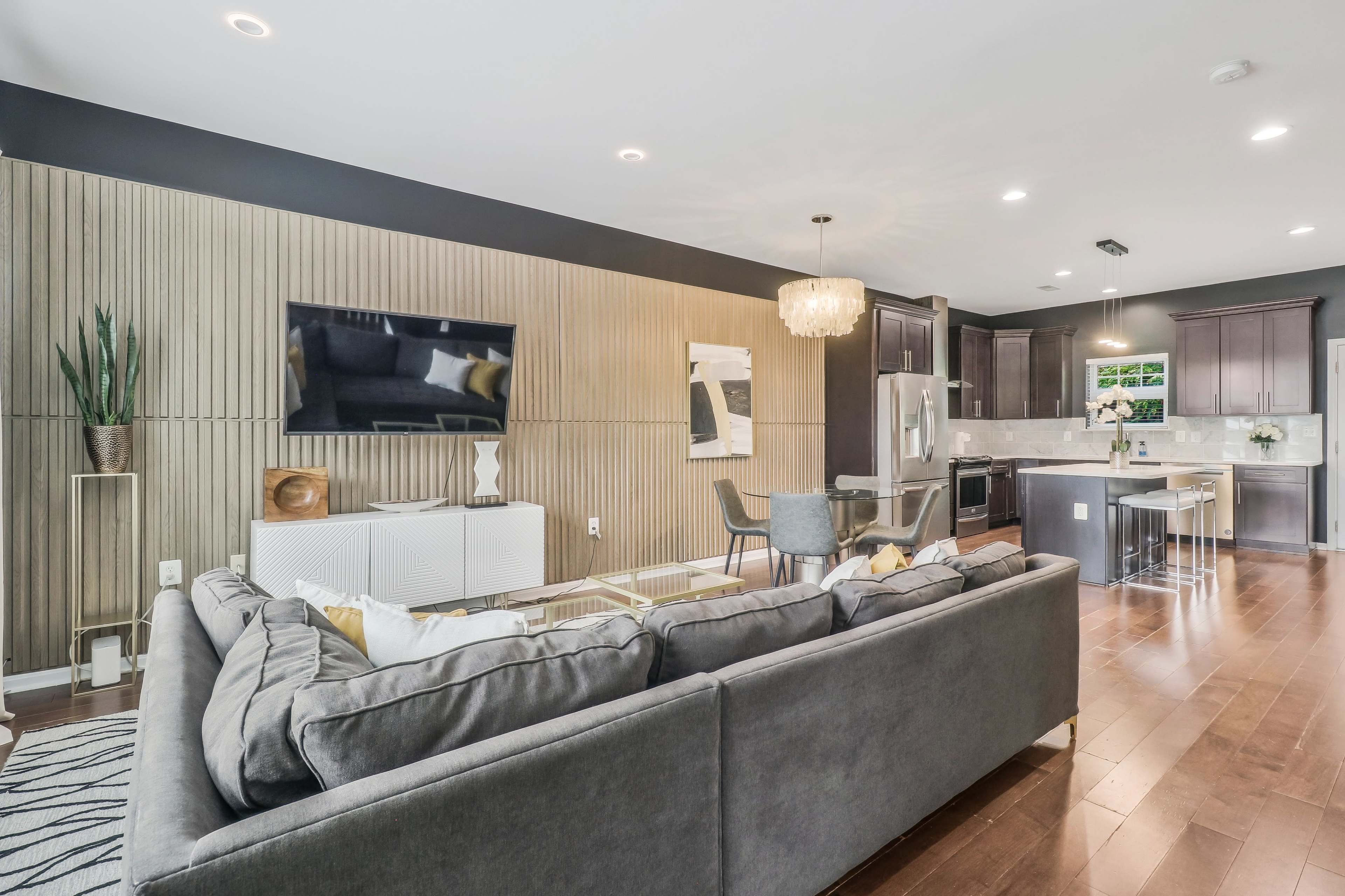 The room features a modern living area with a sectional sofa, a TV mounted on a wood-paneled wall, and an adjacent kitchen with dark cabinetry and a dining table.