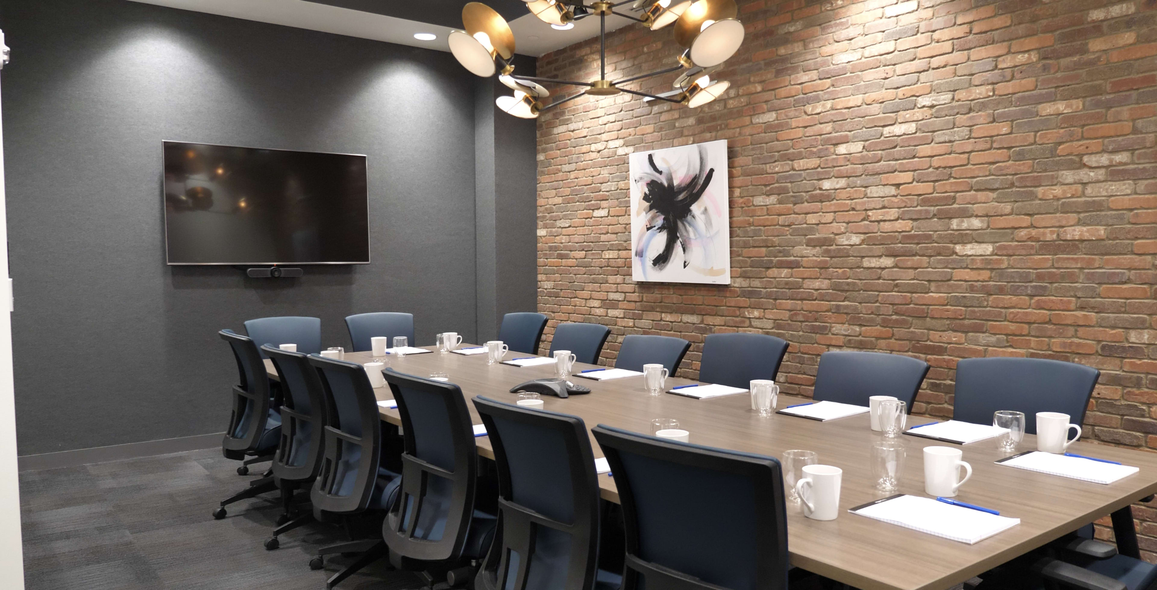A conference room features a long table surrounded by several blue chairs, with a mounted television and a piece of artwork on a brick wall.