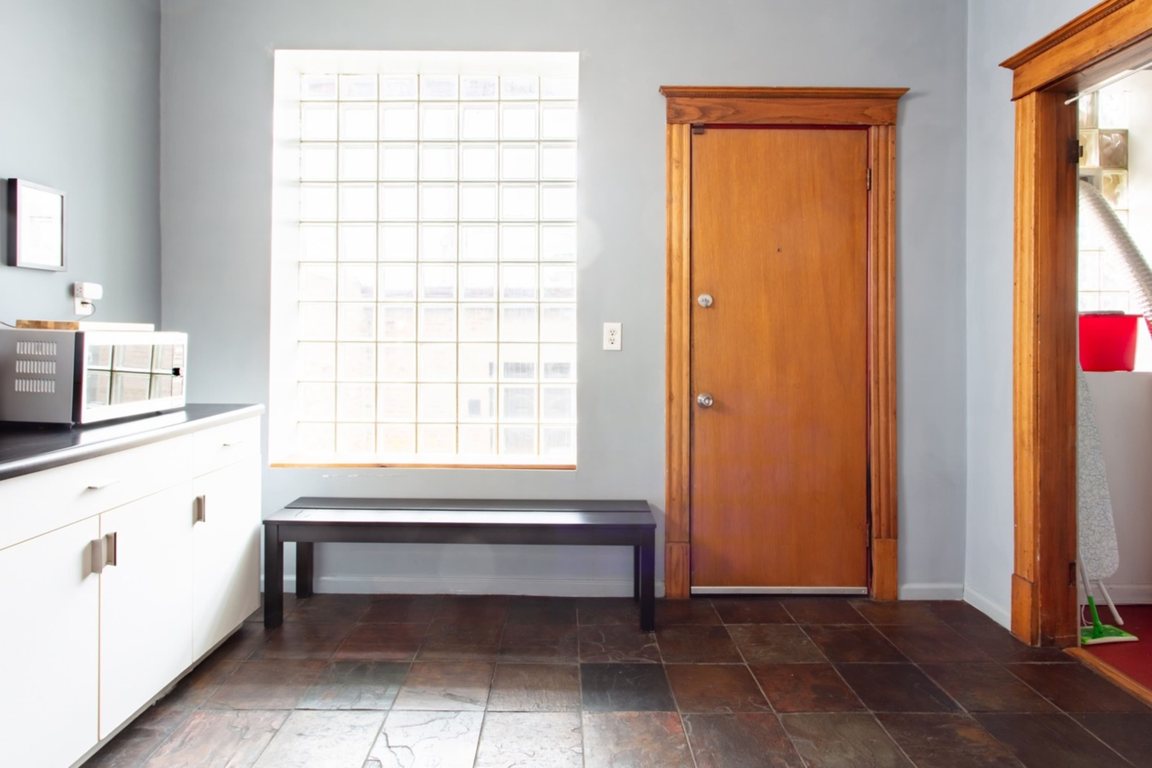 A kitchen area with a wooden door, a large window made of glass blocks, a black bench, and a white cabinet.