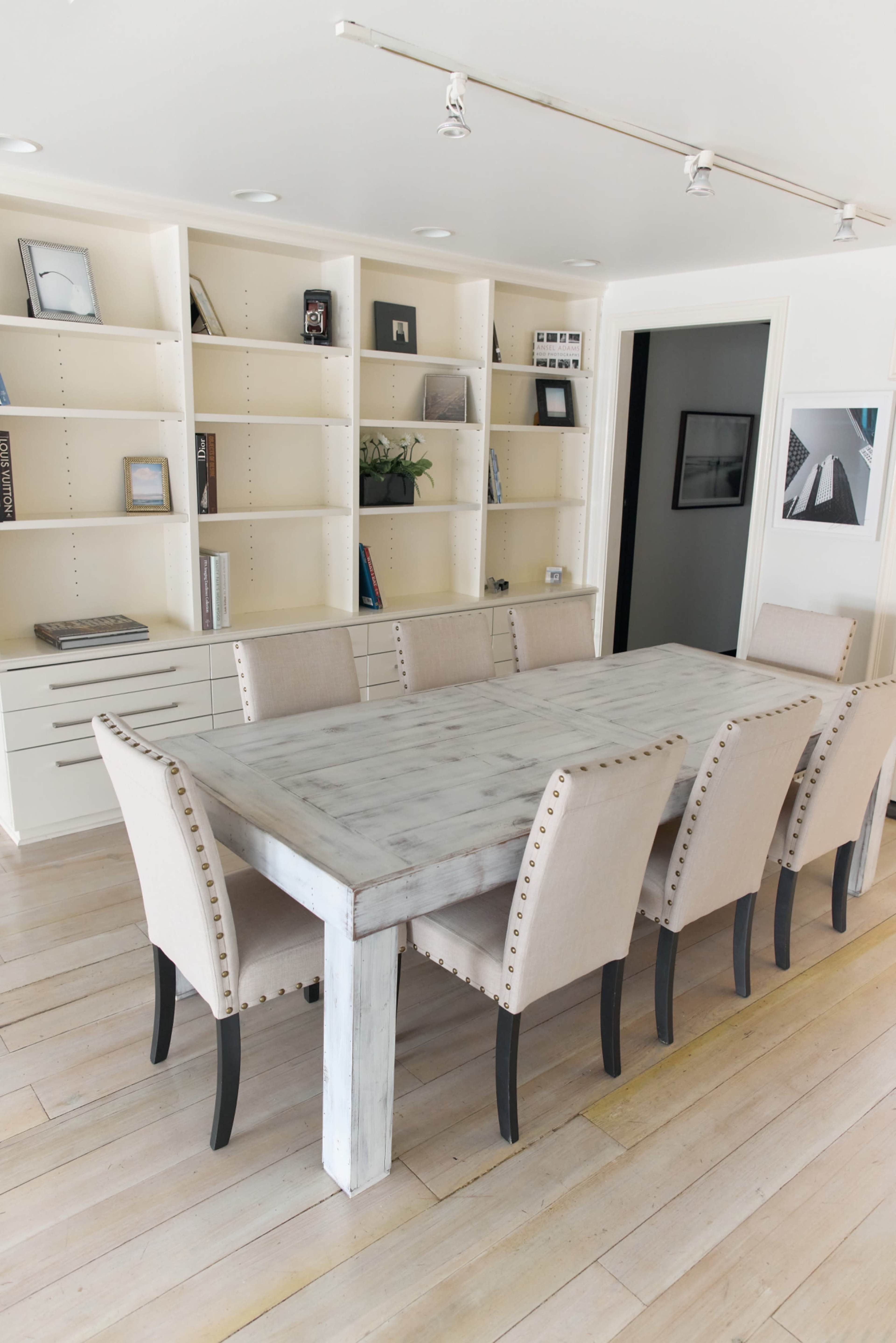 The image shows a modern dining room with a large, rectangular wooden table surrounded by beige upholstered chairs, set against a backdrop of white shelves filled with books and decorative items.