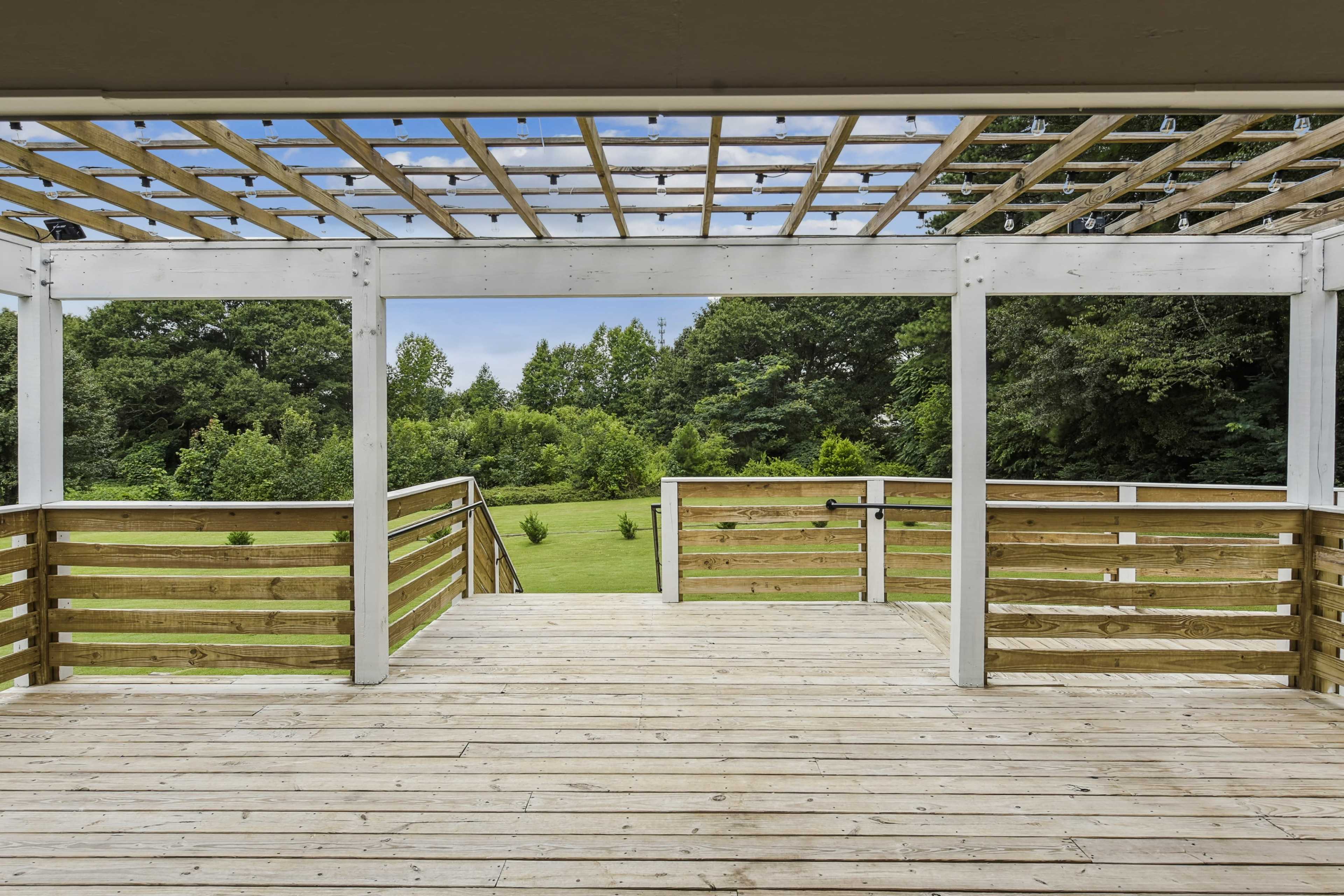 A wooden deck with a lattice roof overlooks a grassy field and wooded area.
