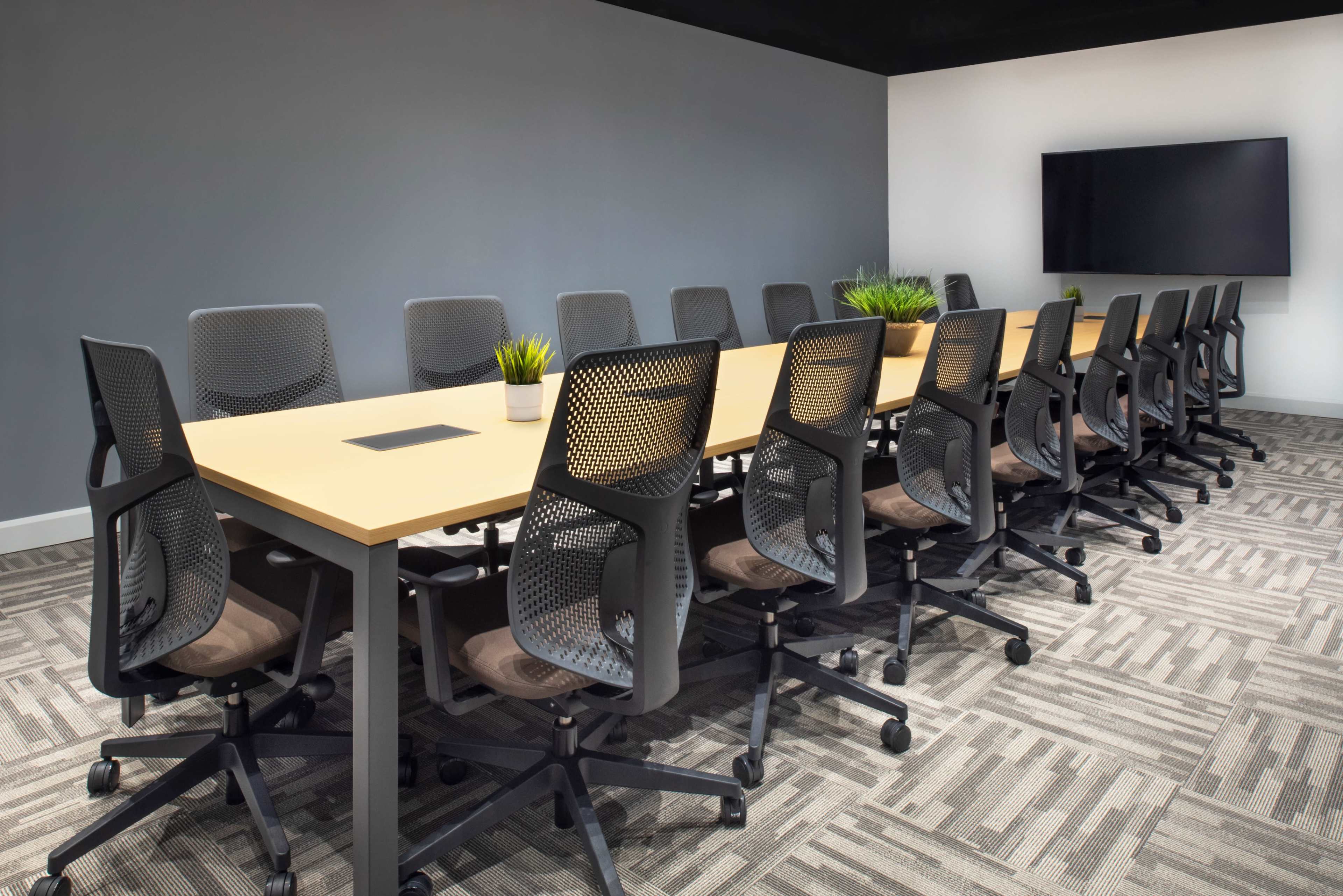 A long conference table is surrounded by black mesh chairs in a modern meeting room with a large screen on the wall.