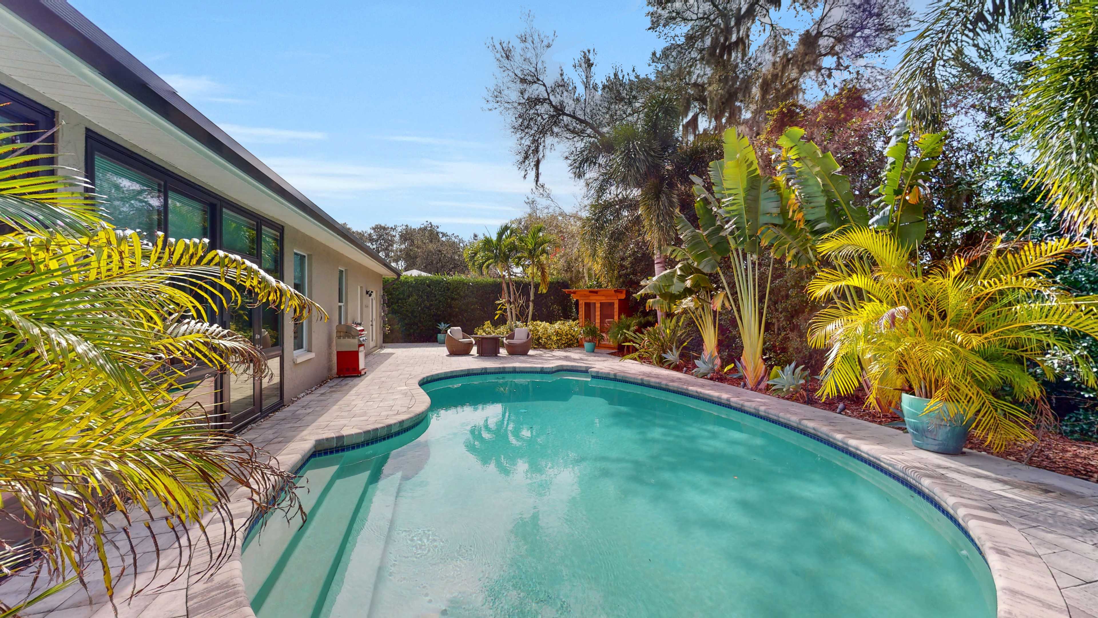 A swimming pool surrounded by tropical plants and a seating area on a sunny day.