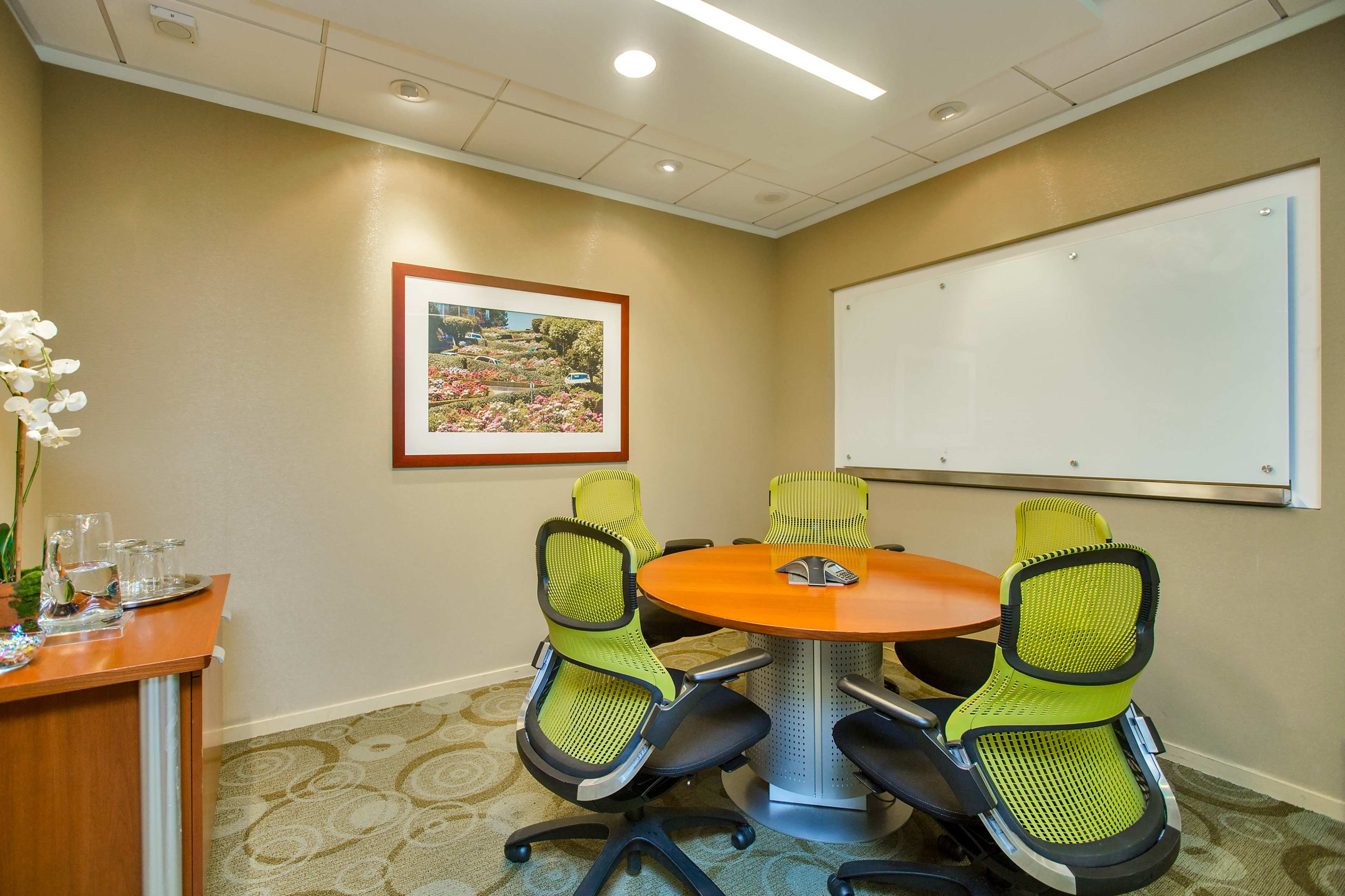 A small meeting room features a circular table surrounded by six green chairs, a framed landscape photograph on the wall, and a whiteboard.