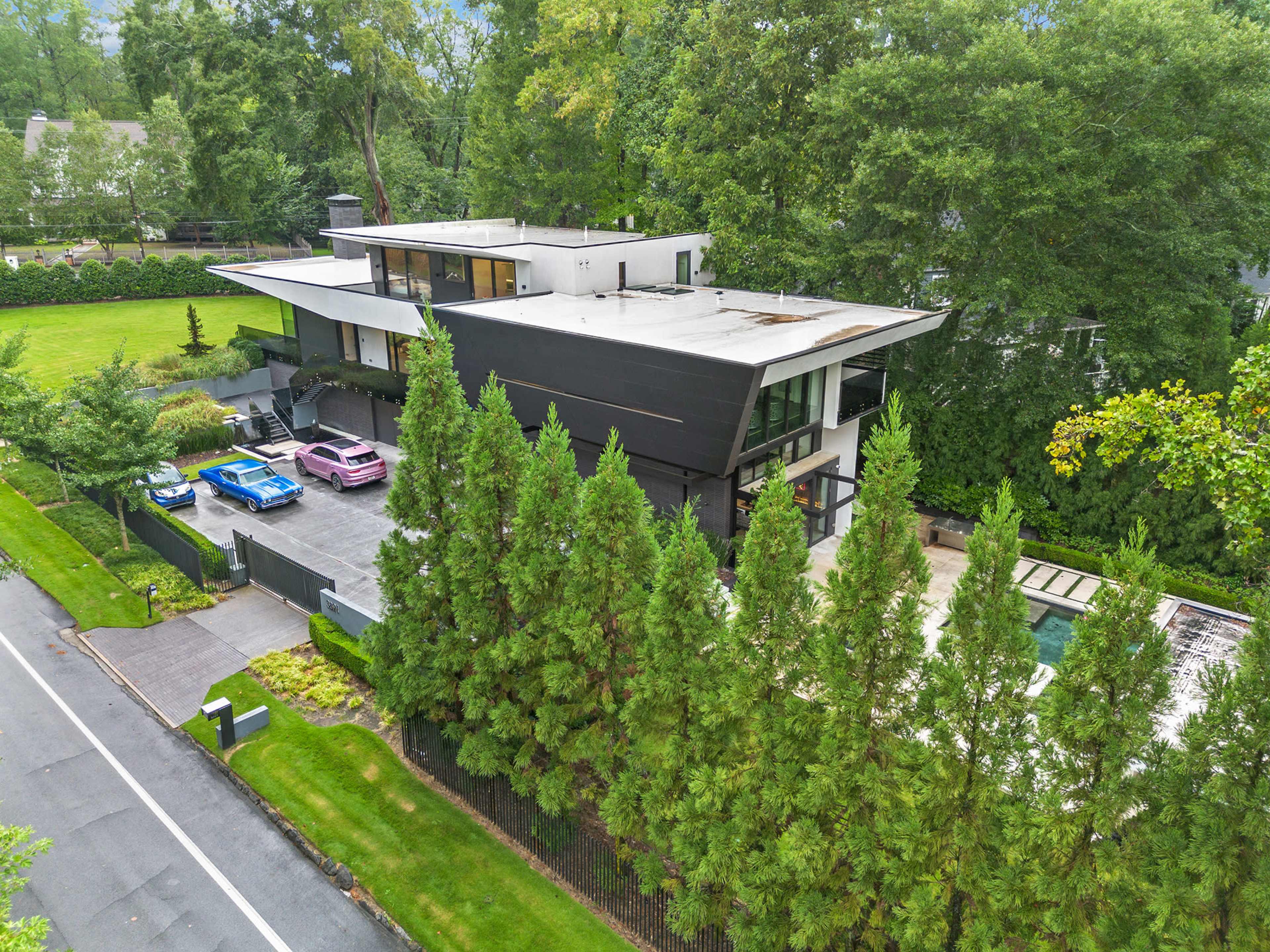 A modern multi-story house with a flat roof and large windows is set among tall green trees, featuring a driveway with several luxury cars parked in front.