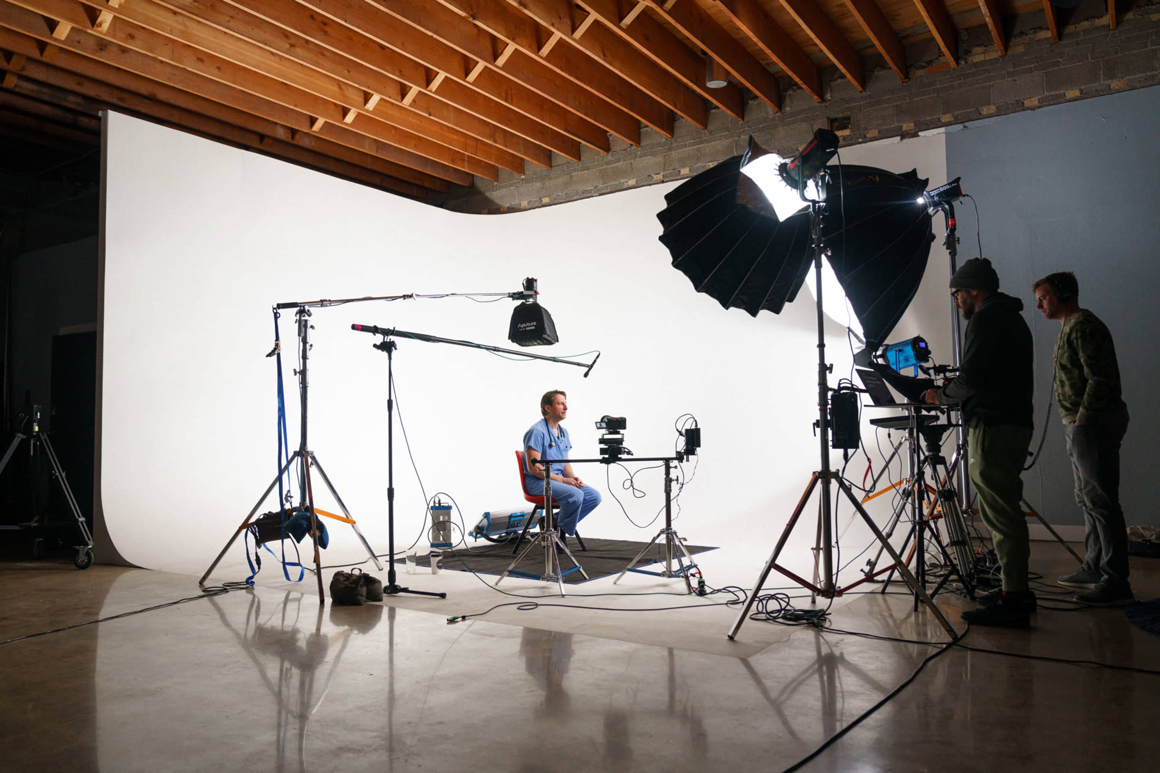 A man sits at a table in front of a white backdrop while a camera and lighting equipment are set up nearby.